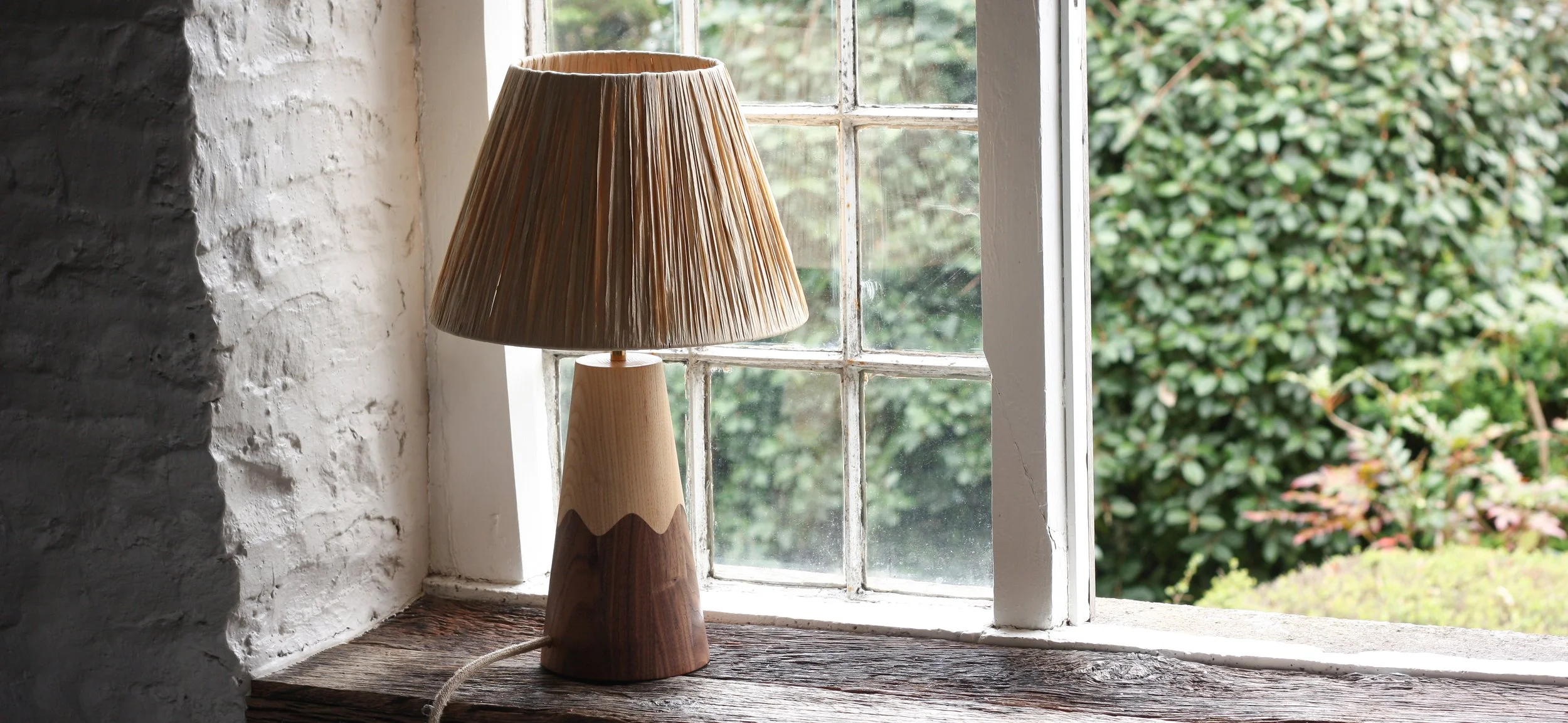 A wooden table lamp with a pleated beige lampshade sitting on a wooden window sill next to a multi pane window with white trim, overlooking green bushes outside.