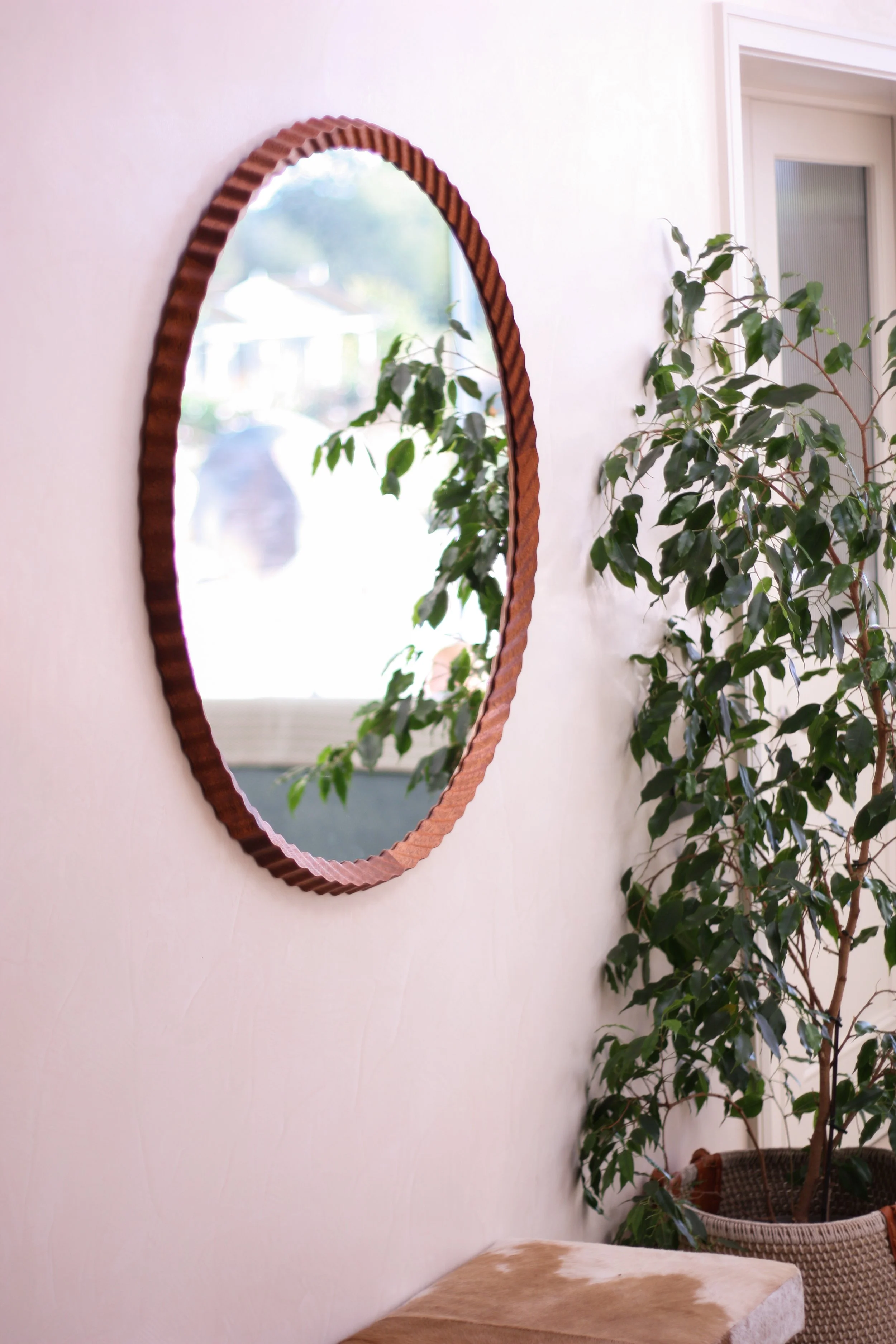 A round, copper-colored mirror hangs on a white wall, reflecting a blurred outdoor scene. To the right, there is a large green leafy plant in a woven basket. In the foreground, part of a beige and brown animal-print upholstered chair or ottoman is visible.