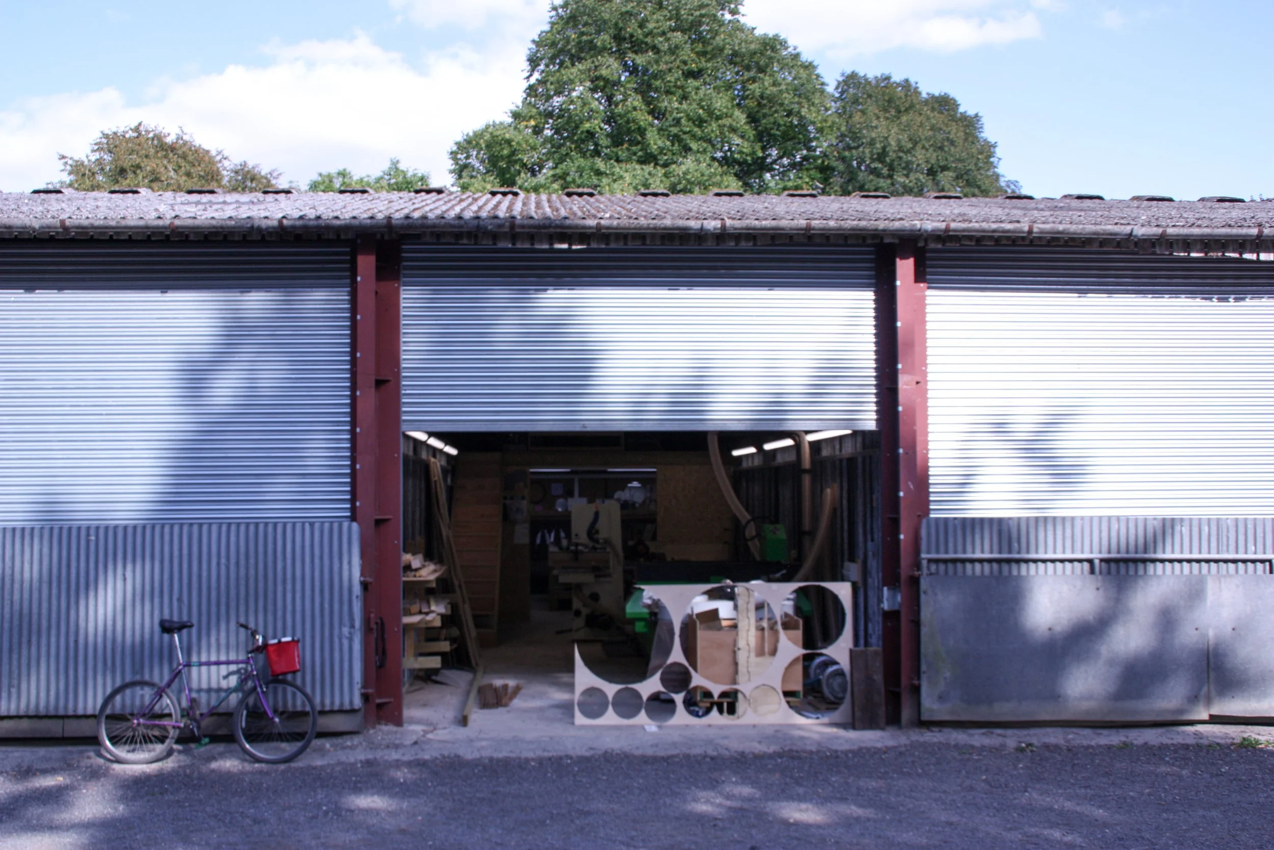 A building with metal siding and a small opening showing inside storage or workshop area, with a bicycle and various wooden or metal objects outside, and trees and sky in the background.