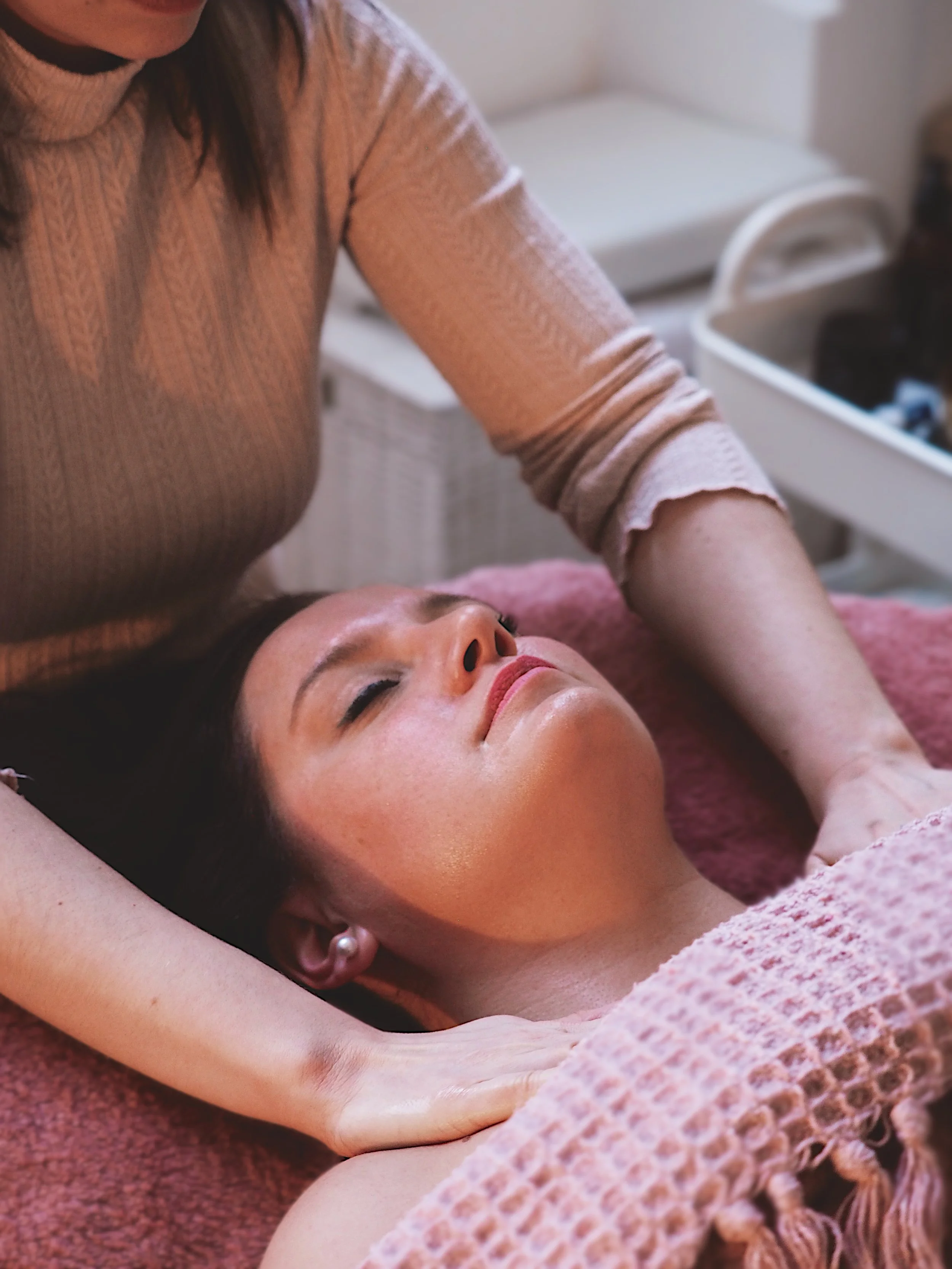 A woman receives a massage on her shoulders while lying on a massage table. She appears relaxed with closed eyes, and a massage therapist is applying pressure to her shoulders.