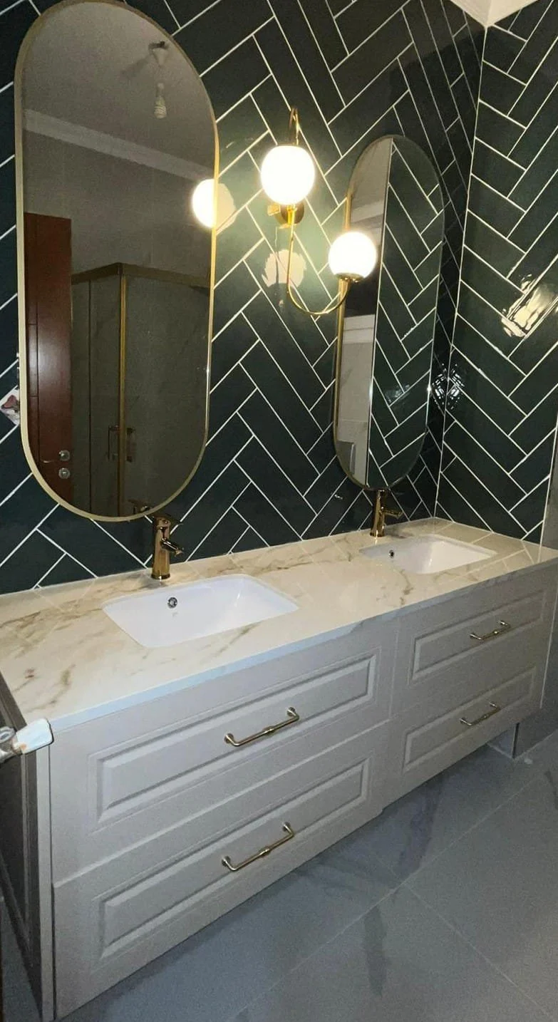Bathroom vanity with two oval mirrors, black tile wall with white grout, gold fixtures, and a beige marble countertop with two sinks.