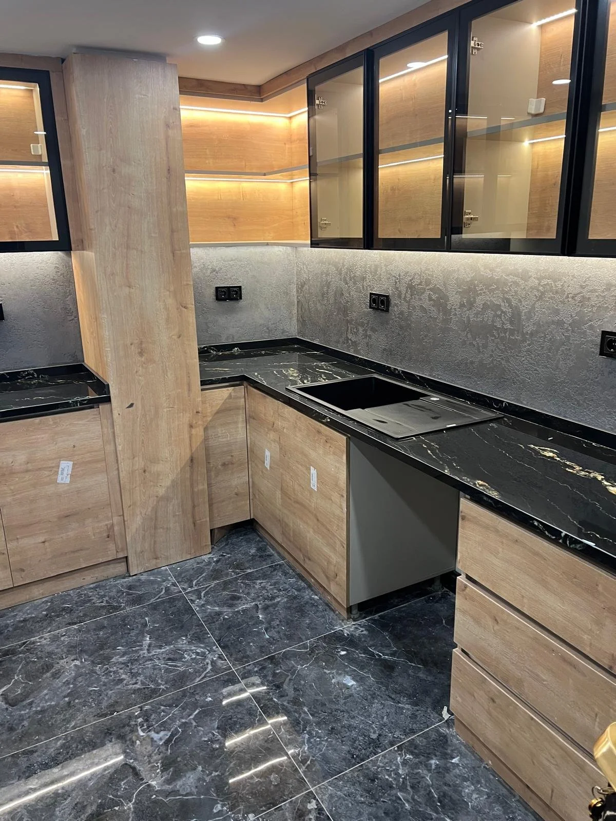 Modern kitchen with black marble countertops, wooden cabinetry, glass upper cabinets, and a black marble floor.
