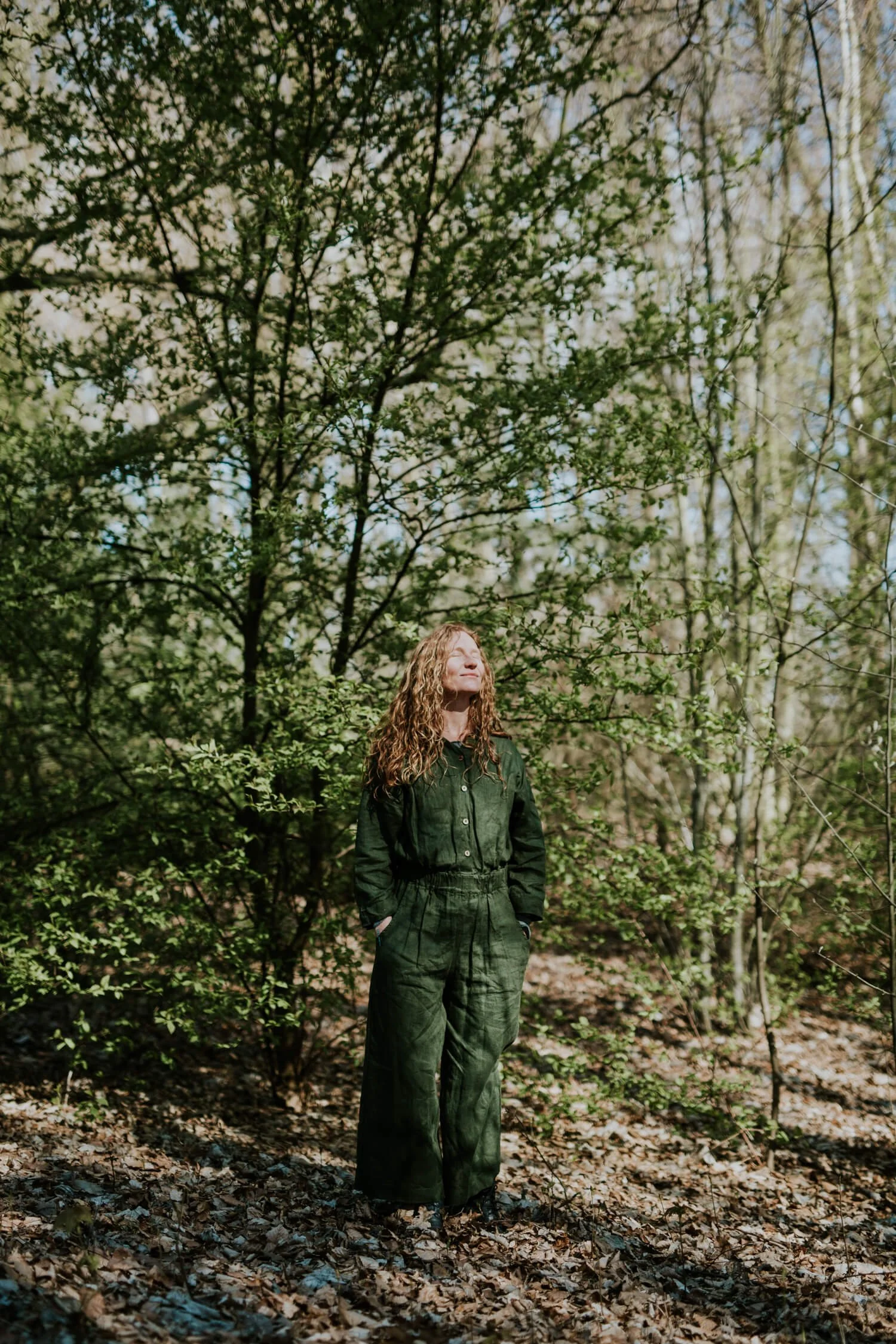 En kvinde med rødt langt krøllet hår iført en grøn heldragt står med lukkede øjne og ansigtet vendt mod solen foran et nyudsprunget lille bøgetræ