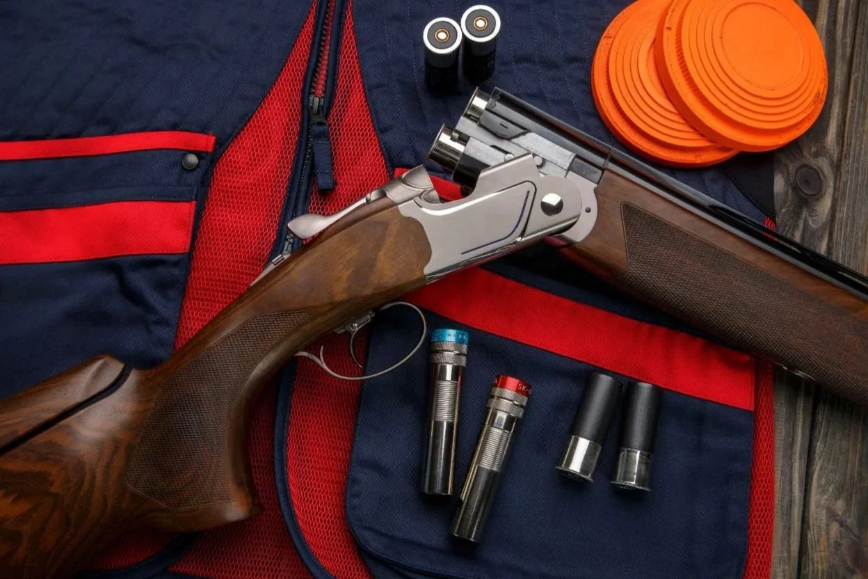 A man and a woman at a shooting range, with the man instructing the woman as she aims a shotgun outdoors.