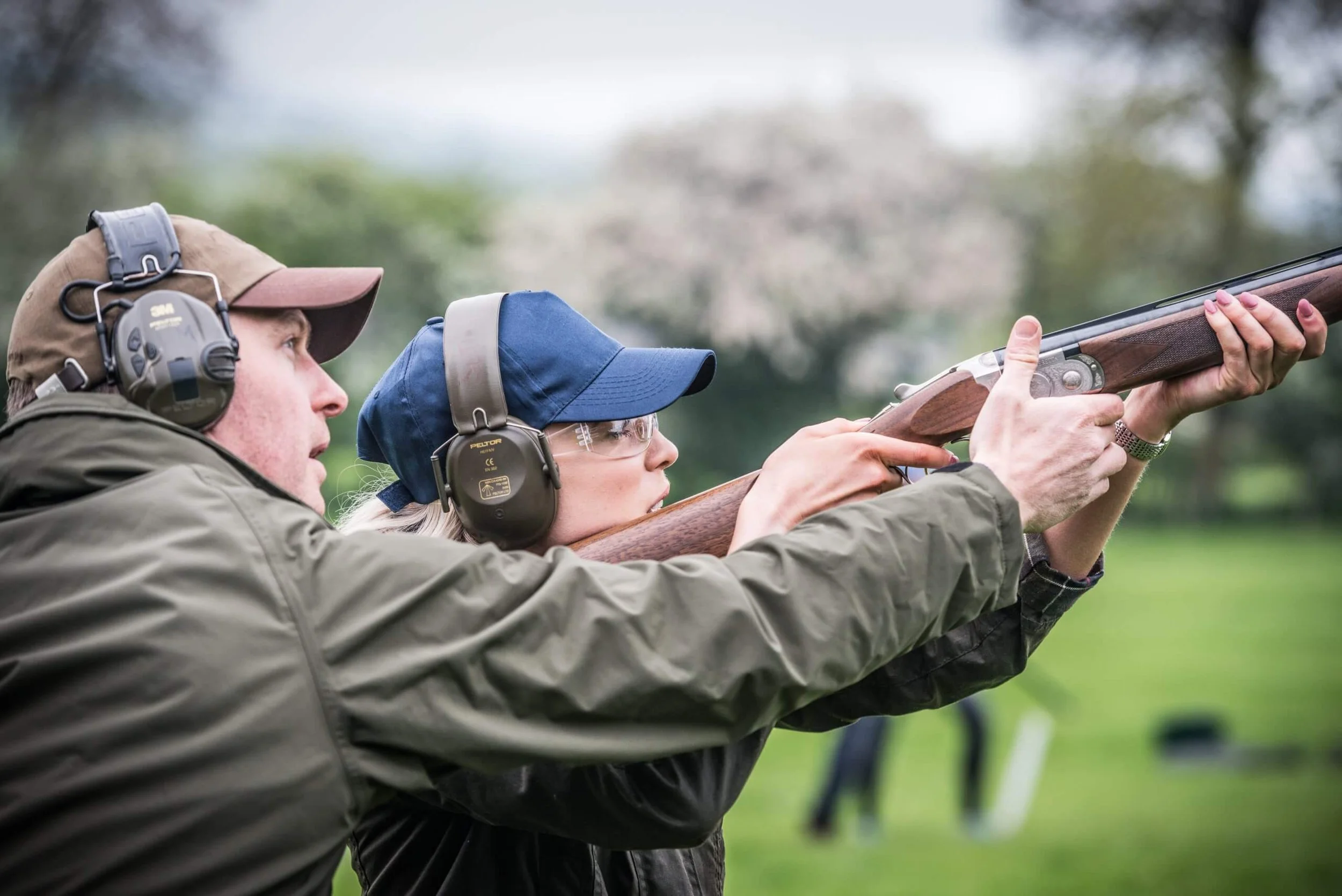 A man and a woman at a shooting range, with the man instructing the woman as she aims a shotgun outdoors.