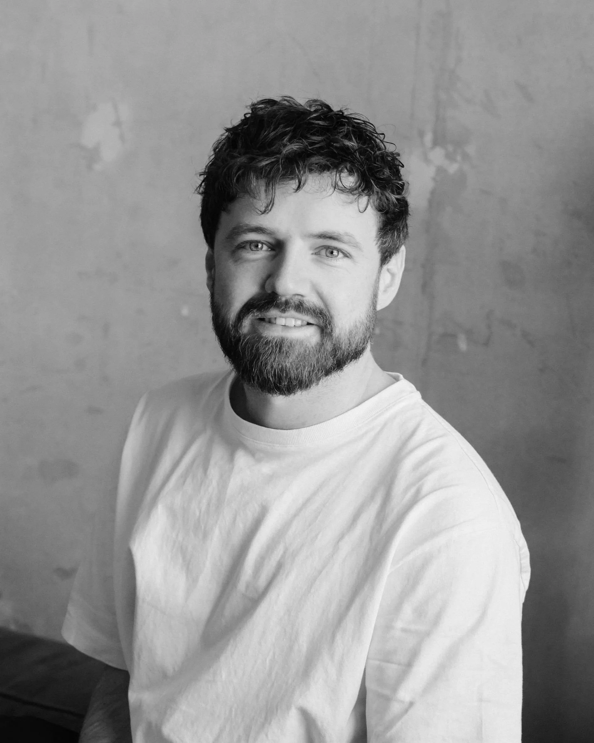 Black and white photo of a man with curly hair and a beard, smiling, wearing a white shirt, against a plain wall background.