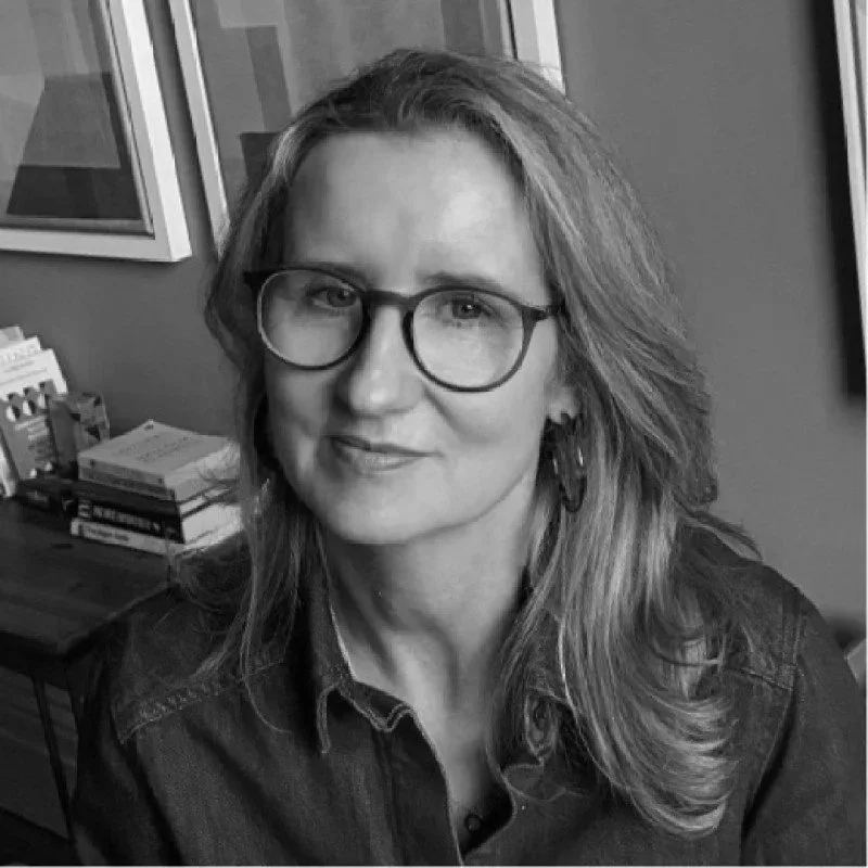 Black and white photo of a middle-aged woman with free-flowing wavy hair, wearing glasses, a denim shirt, and earrings, sitting in a room with framed pictures on the wall and a table with books and papers.