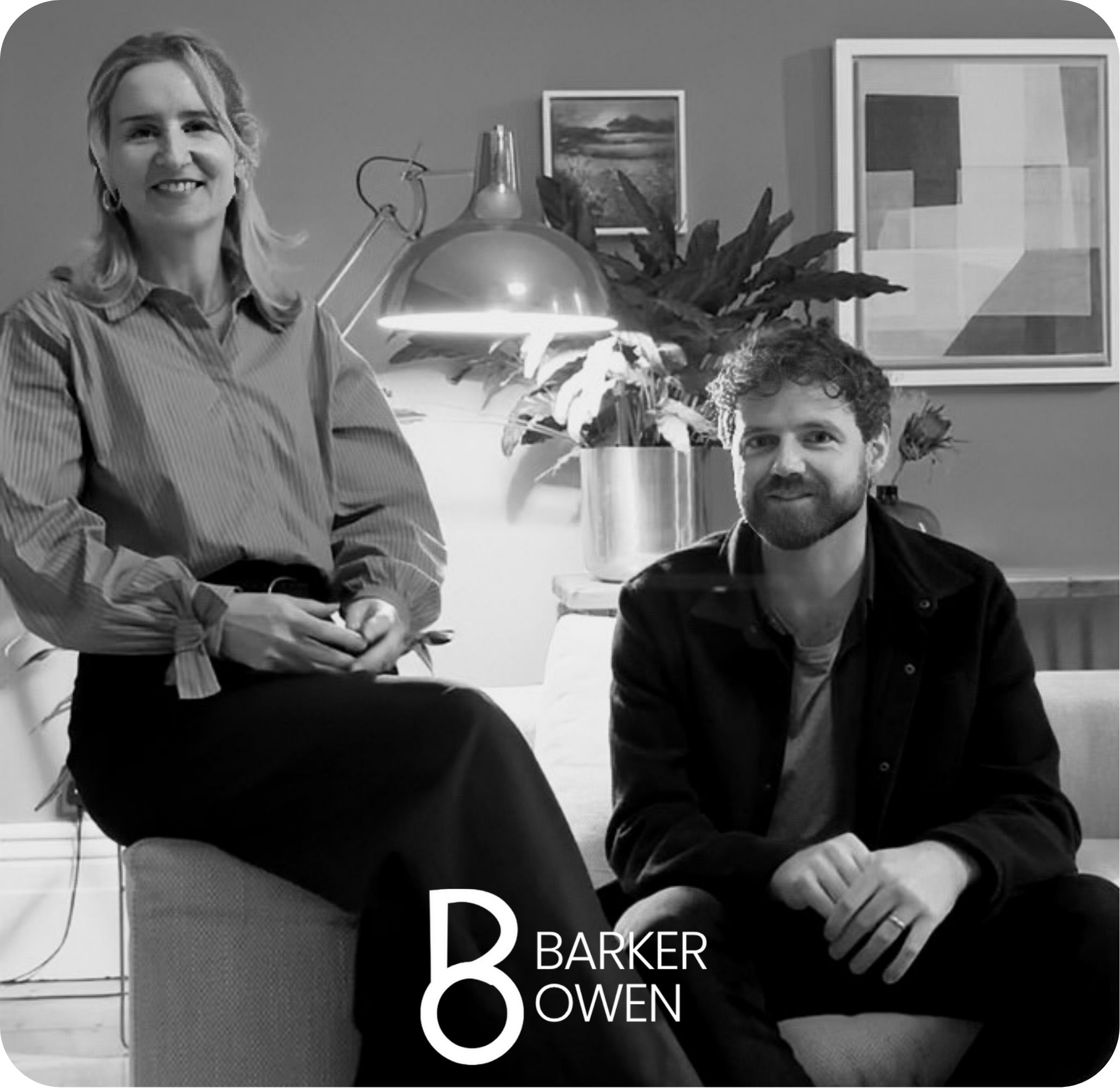 A woman sitting on a sofa and a man sitting on a chair next to her in a living room with modern art and large potted plant in the background.