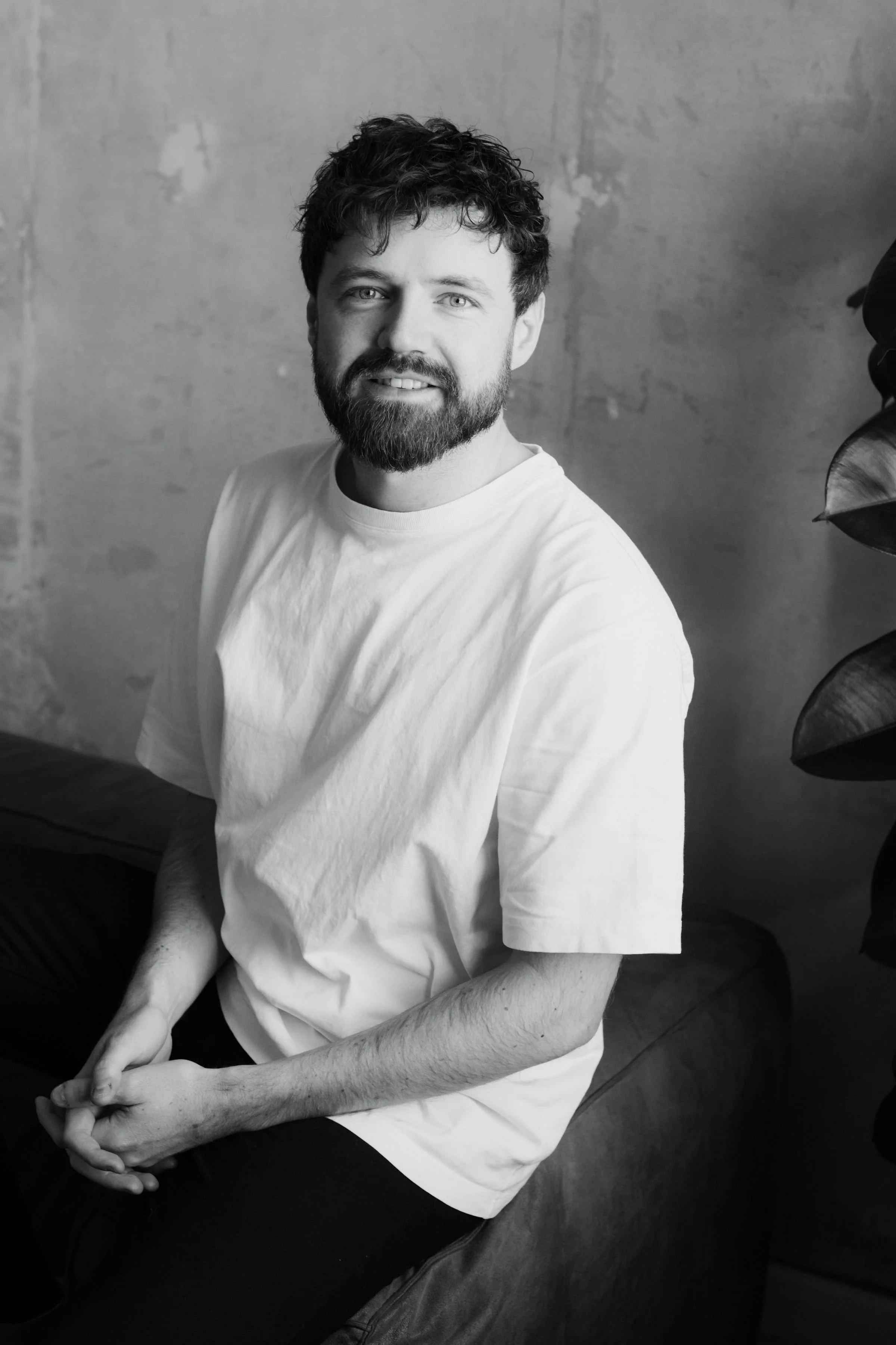 Black and white photo of a bearded man with curly hair, wearing a plain light-colored T-shirt, sitting on a bench with a plain wall in the background, and large plant leaves partially visible on the right side.