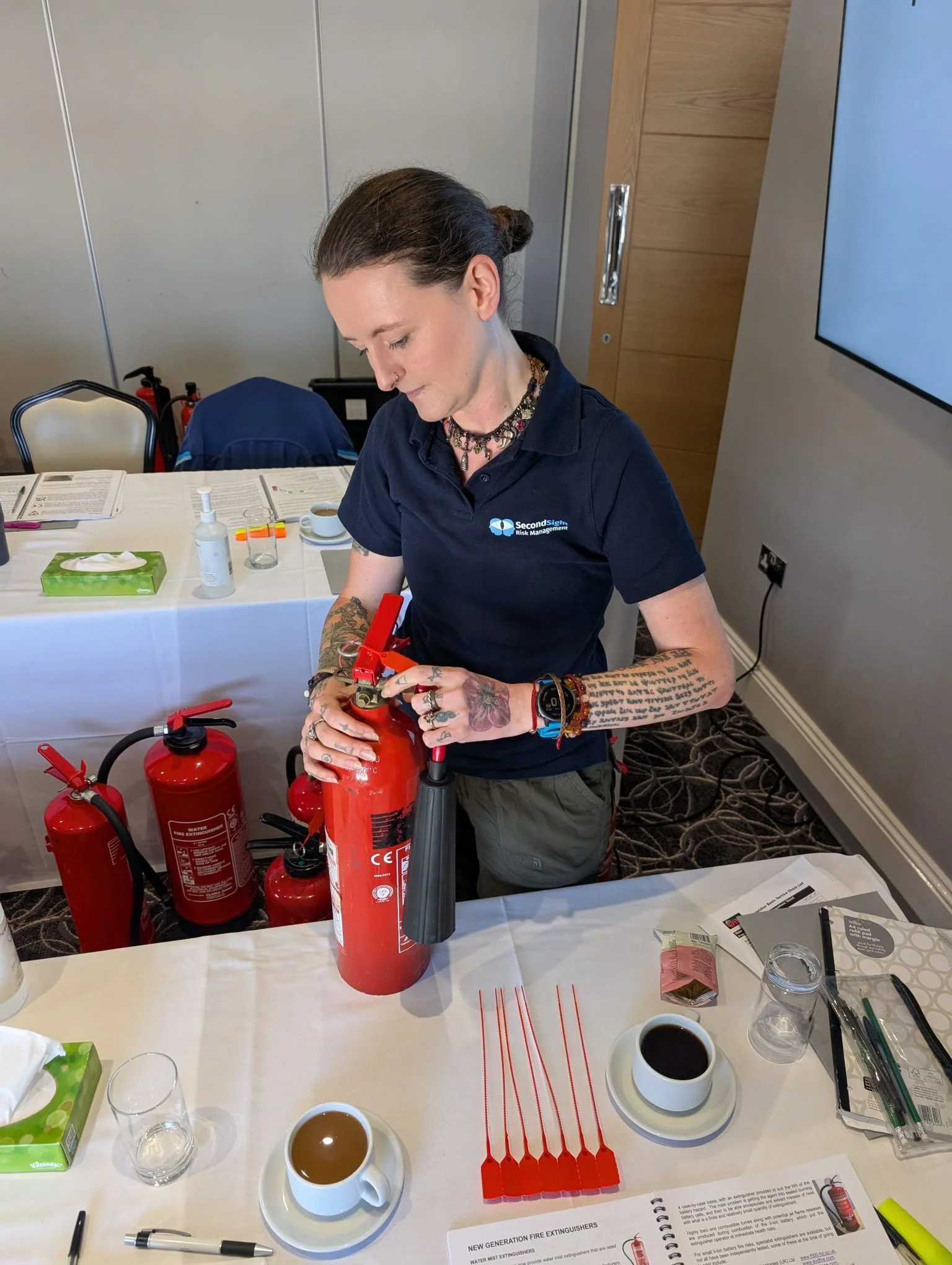 A woman with tattoos on her arms and wearing a black polo shirt with a logo, standing at a table with red fire extinguishers, cups of coffee, and documents. She is inspecting or working on a fire extinguisher in an indoor setting.