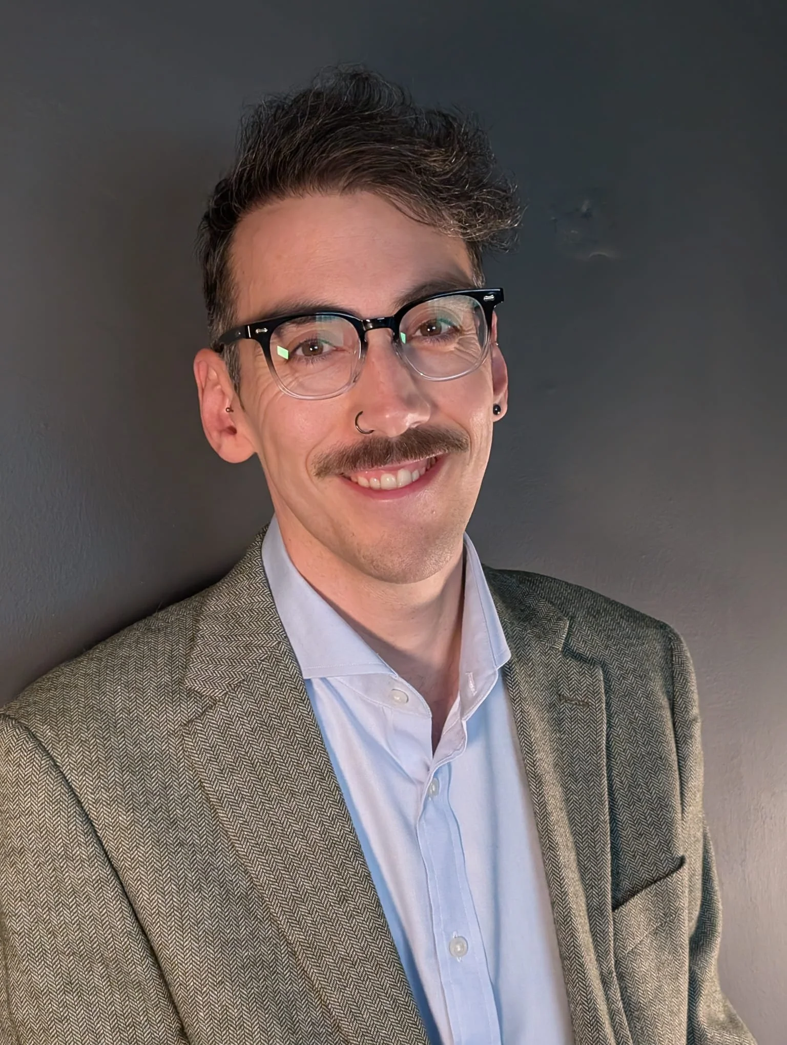 A man with glasses, a mustache, and short, dark, curly hair smiling, wearing a blazer and white shirt, standing against a dark gray background.