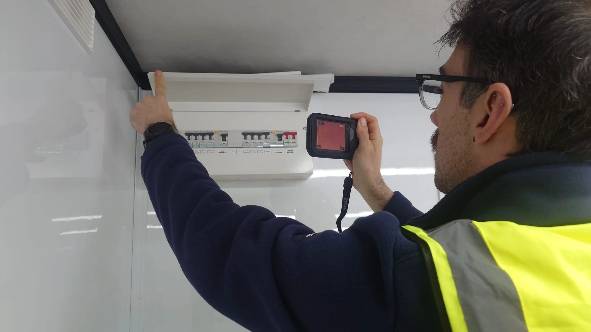A man wearing glasses and a reflective safety vest is inspecting an electrical panel or circuit breaker box, pointing at one of the circuit breakers while holding a thermal imaging camera.