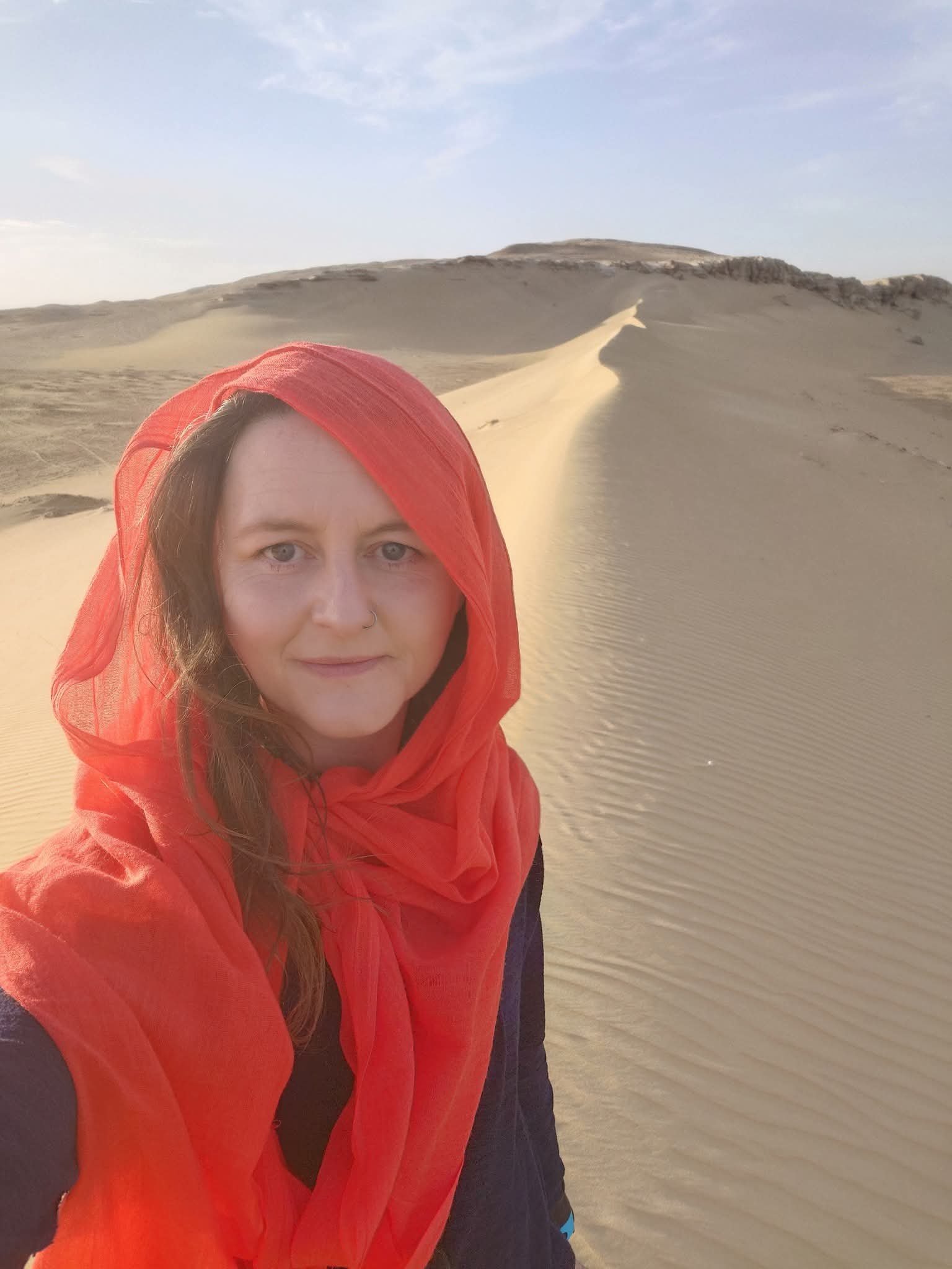 A woman with a red head covering and black jacket standing in a sand desert with dunes and a blue sky.