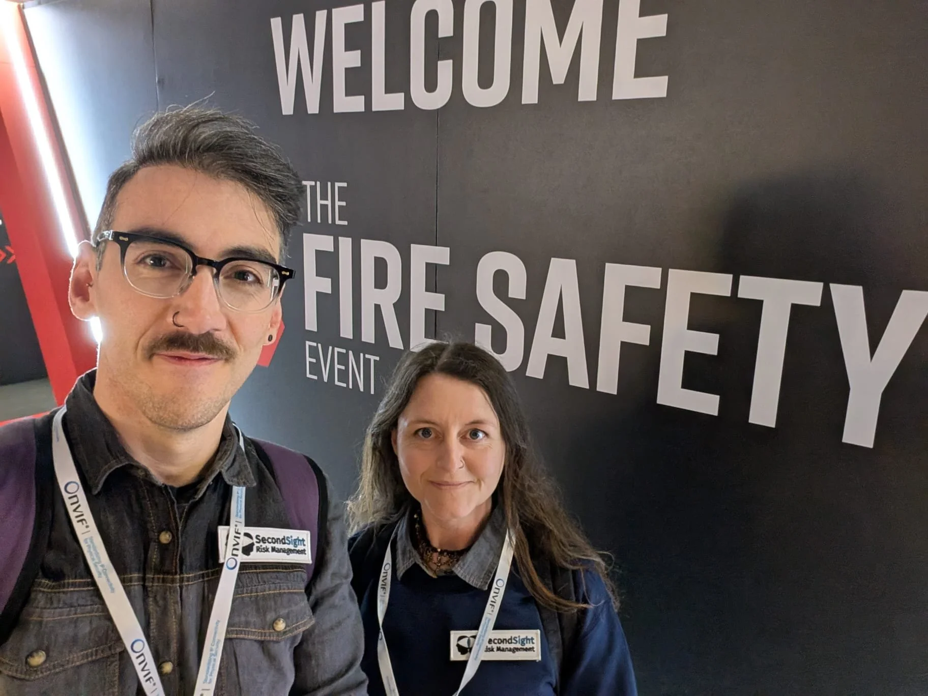 Two people, a man and a woman, taking a selfie in front of a black wall with bold white text that says 'WELCOME TO THE FIRE SAFETY EVENT.' They are wearing conference badges on lanyards around their necks.