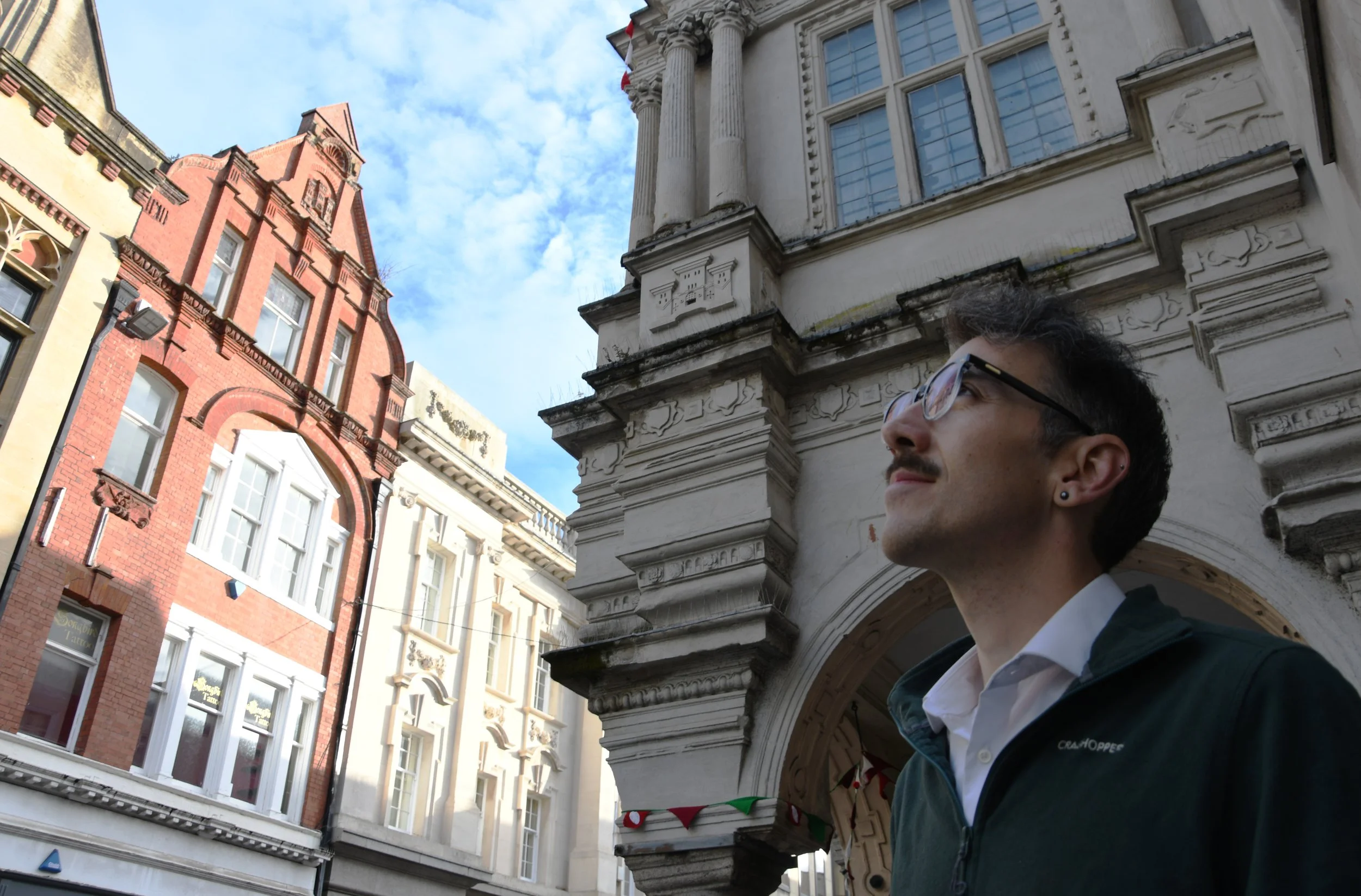 A man with glasses and a mustache standing outdoors in front of historic buildings with ornate architecture, looking upward.
