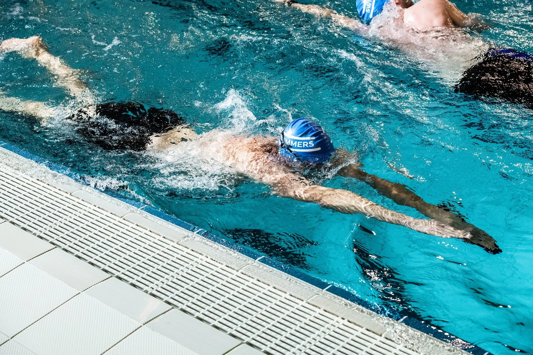 Een persoon zwemt in een binnenzwembad met een blauwe badmuts met de tekst 'SWIMMERS'