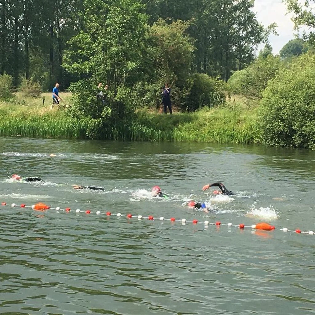 Mensen zwemmen in een open water zwemwedstrijd met een gele lijn met oranje en witte drijvers in een groene omgeving met bomen.