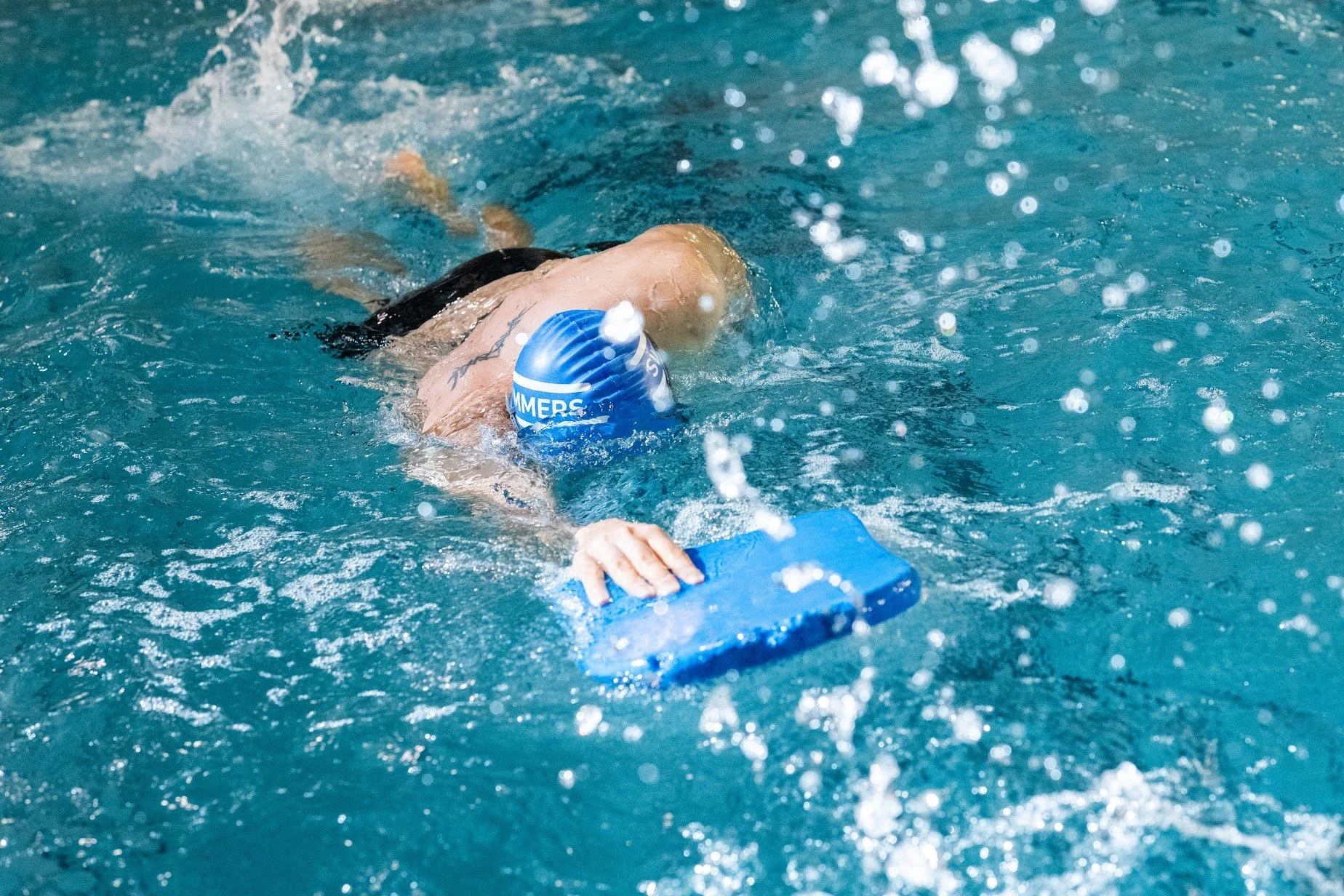 Een zwemmer met een blauwe cap en zwembril rust aan de rand van een binnenzwembad.