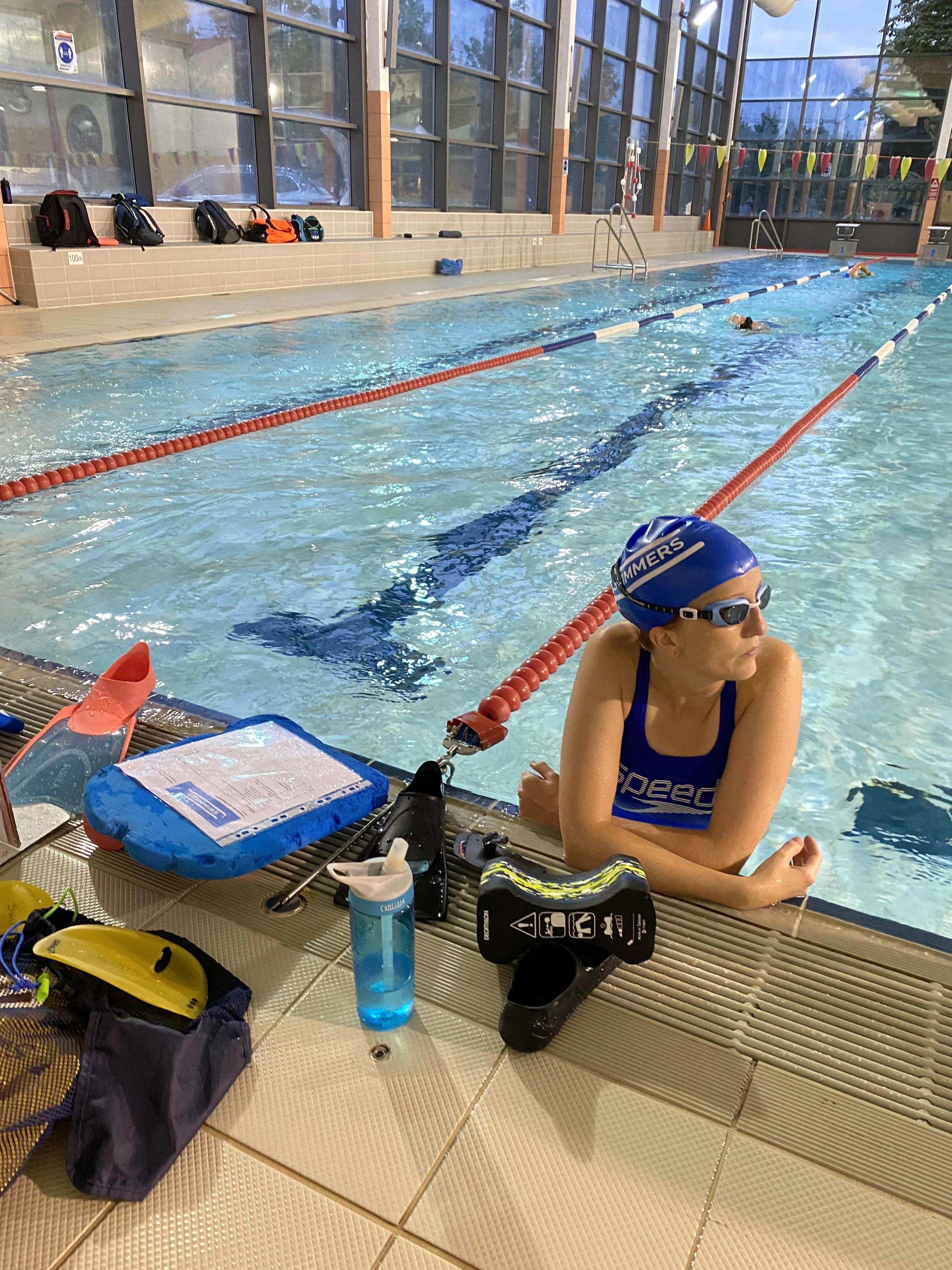 Een vrouw in een zwembril en badpak aan de rand van het zwembad, klaar om te zwemmen, met zwemuitrusting zoals zwemvoer, stopwatch en waterfles op de rand.