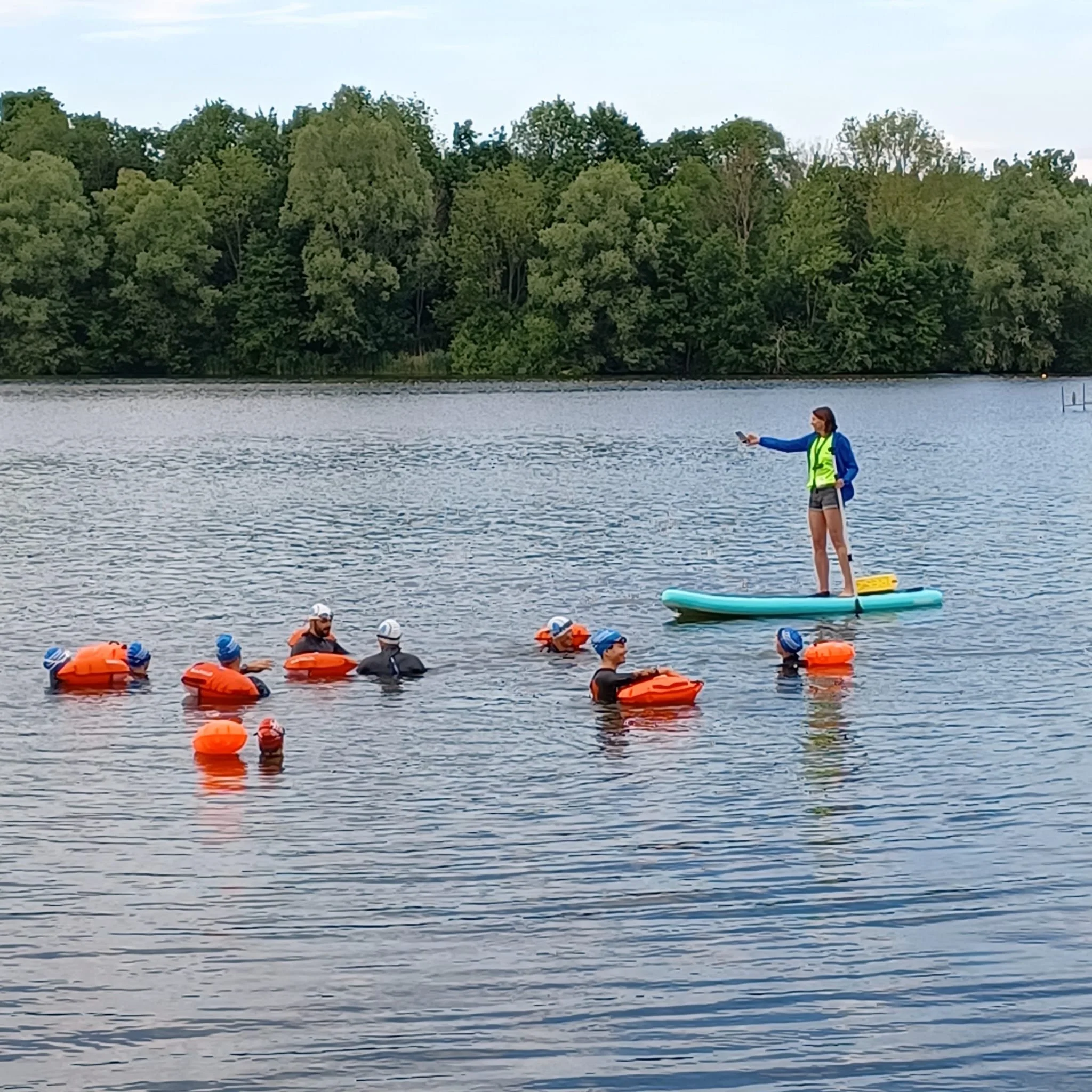 Een instructeur op een paddleboard geeft les aan een groep zwemmers met orange zwembrillen en zwemvliezen in een meer, omringd door groene bomen.
