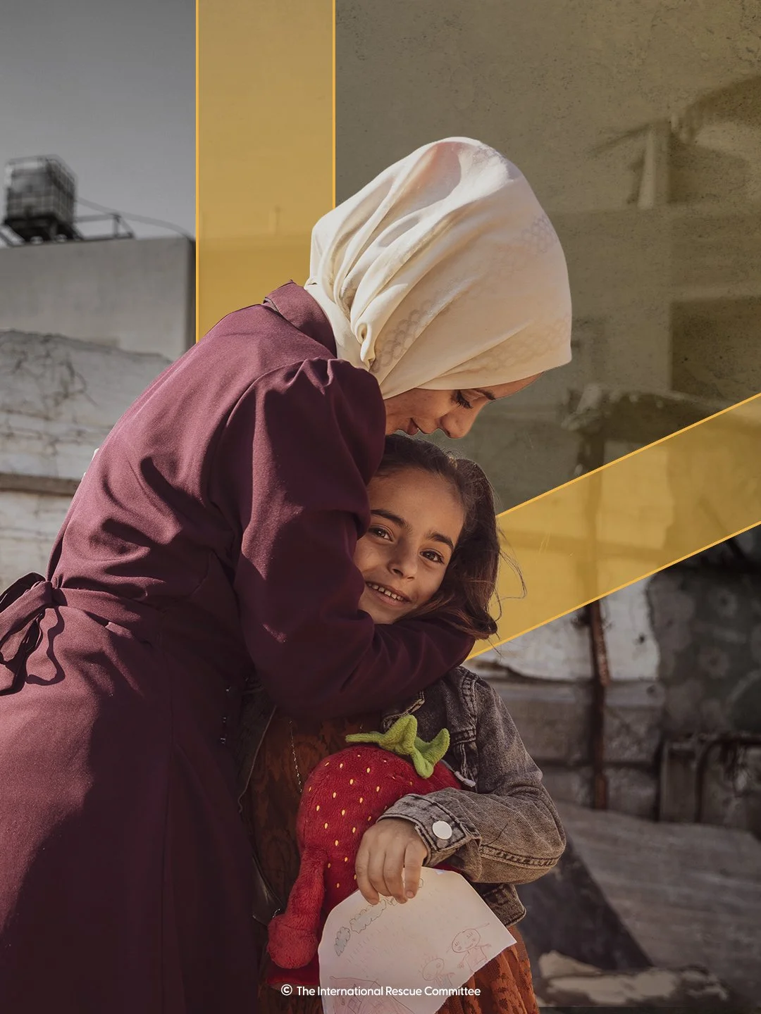 A woman with a hijab embracing a smiling young girl holding a stuffed strawberry toy and a drawing. The background shows an urban setting with buildings and a cloudy sky.