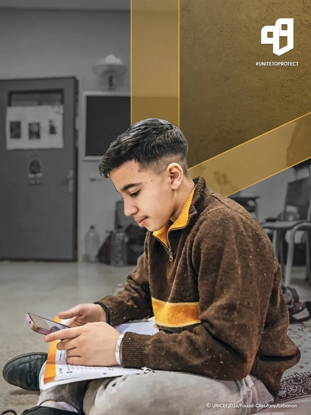 A young boy sitting on the floor in an indoor setting, looking at his smartphone and holding a book or notebook, with a backpack and a water bottle nearby.
