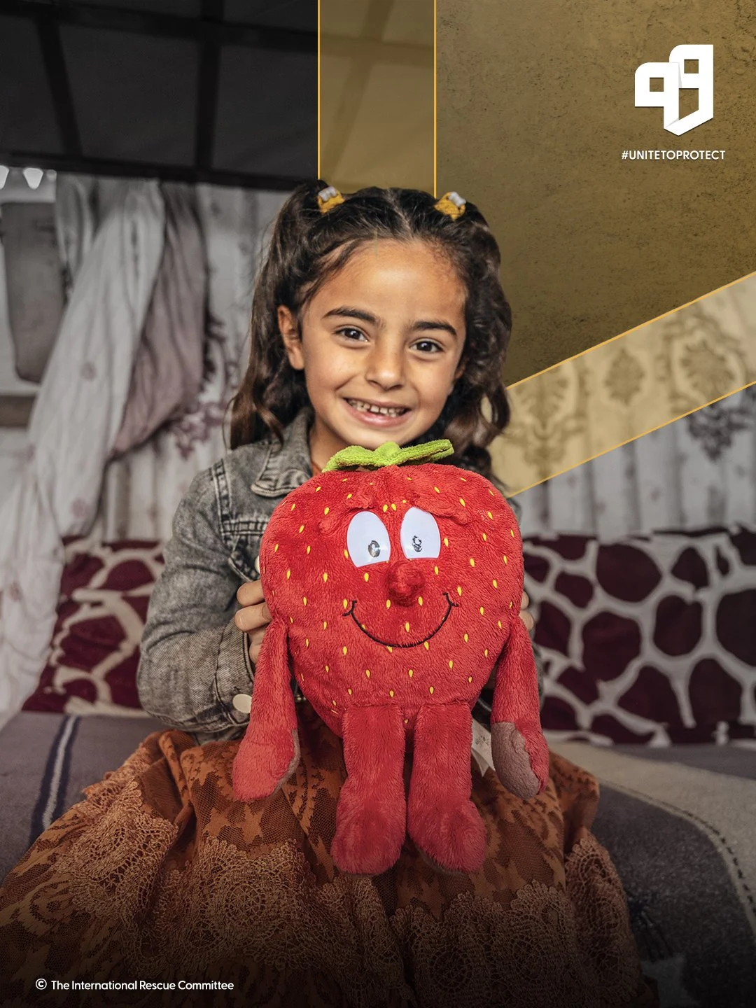Young girl smiling and holding a plush toy shaped like a smiling strawberry with eyes and a green leaf on top, sitting on a bed in a cozy room.