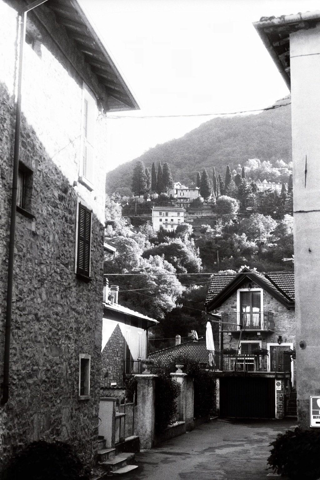 Schmale Straße zwischen alten Stein- und Putzhäusern, im Hintergrund Hügel mit Häusern und Bäumen, im Vordergrund eine Terrasse mit Stühlen und einer Markise.