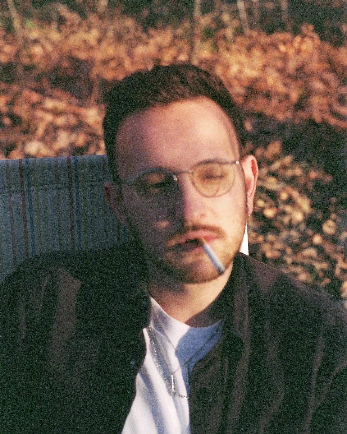 Ein junger Mann mit dunklem Haar, Brille mit gelben Gläsern, trägt eine schwarze Jacke und eine weiße T-Shirt, sitzt draußen vor einem Hintergrund aus Herbstblättern, raucht eine Zigarette und hat eine entspannte Haltung.