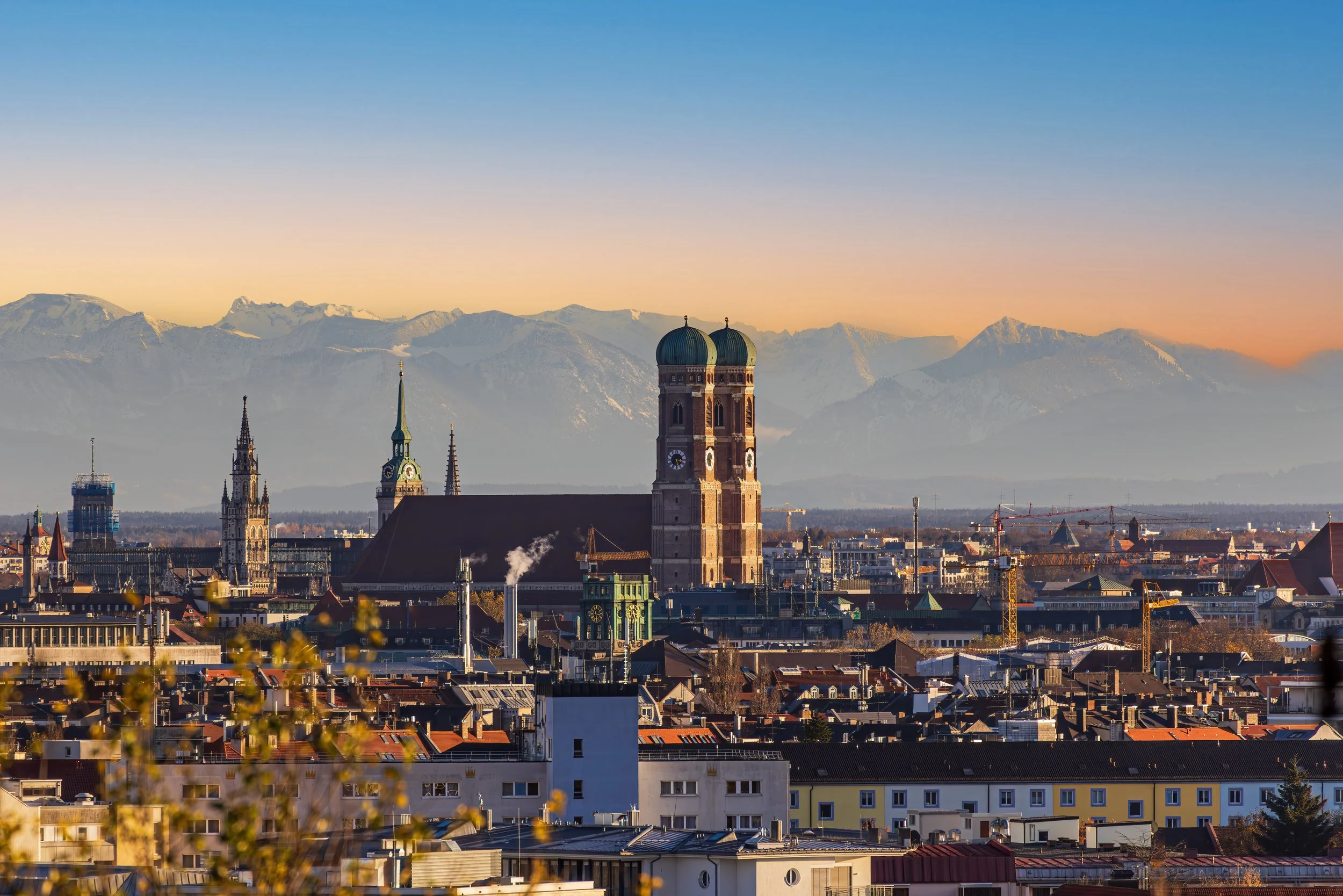 Stadtansicht von München mit Kirchtürme der Frauenkirche, im Hintergrund Alpen bei Sonnenaufgang mit Mehrfamilienhäuser; Immobilienverwaltung München Alexandra Mayr