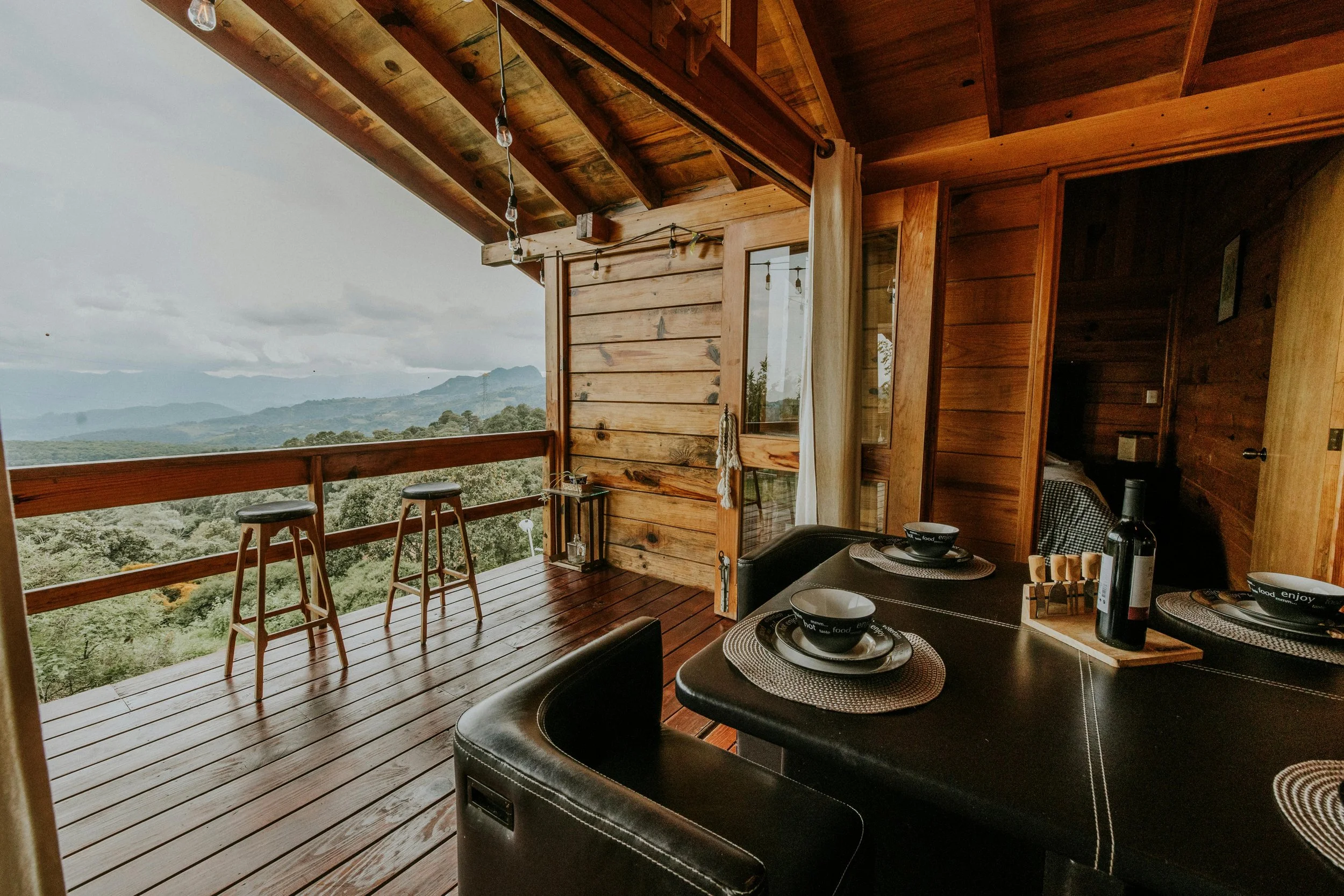Balcon en bois dans une maison en bois avec vue sur des montagnes et la forêt, avec une table à manger et des assiettes à l'intérieur.