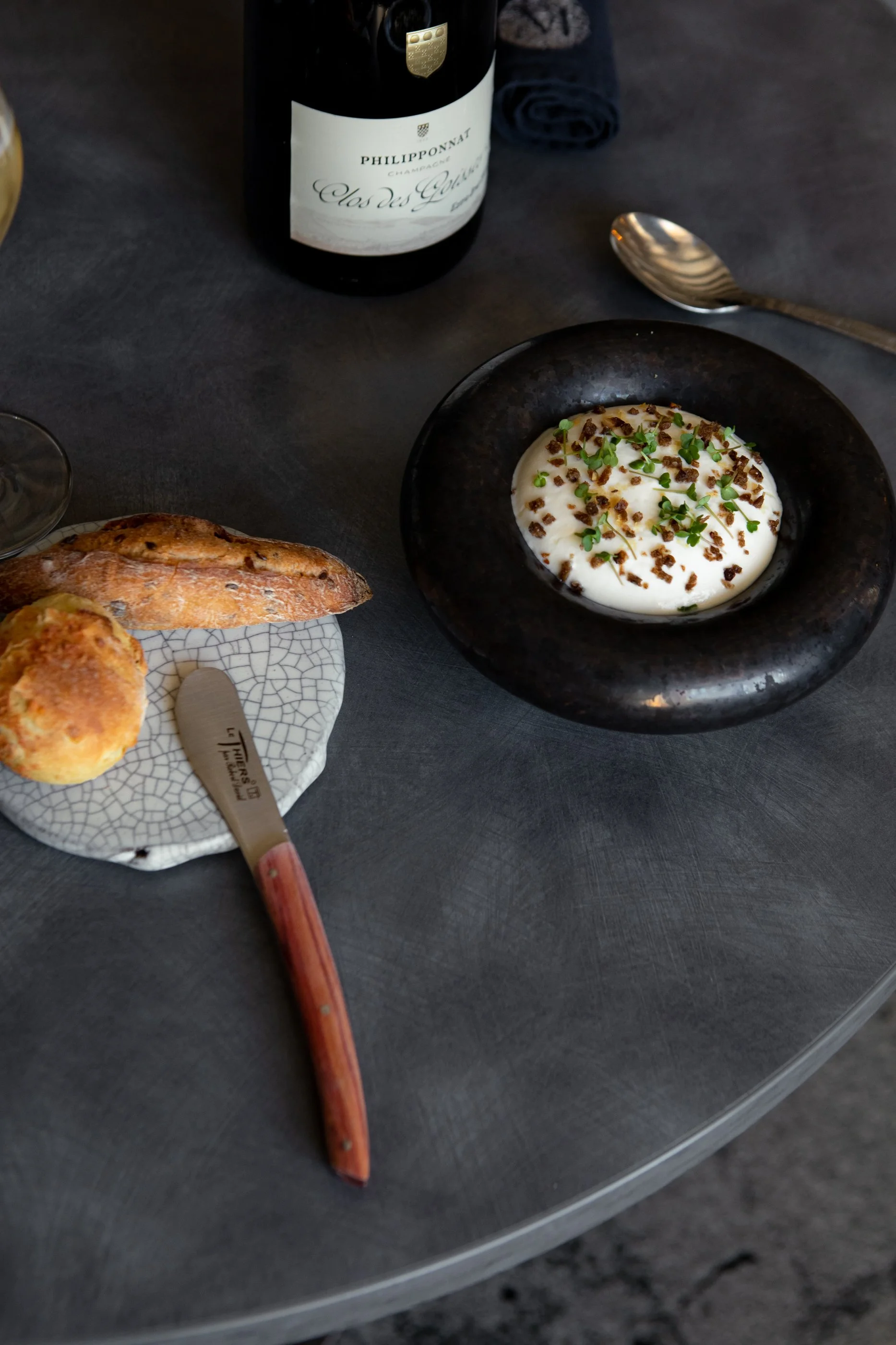 Plat avec un œuf à la coque garni d'herbes et de petits morceaux, accompagnée de pain et de petits pains, avec une bouteille de champagne et une cuillère argentée sur une table sombre.