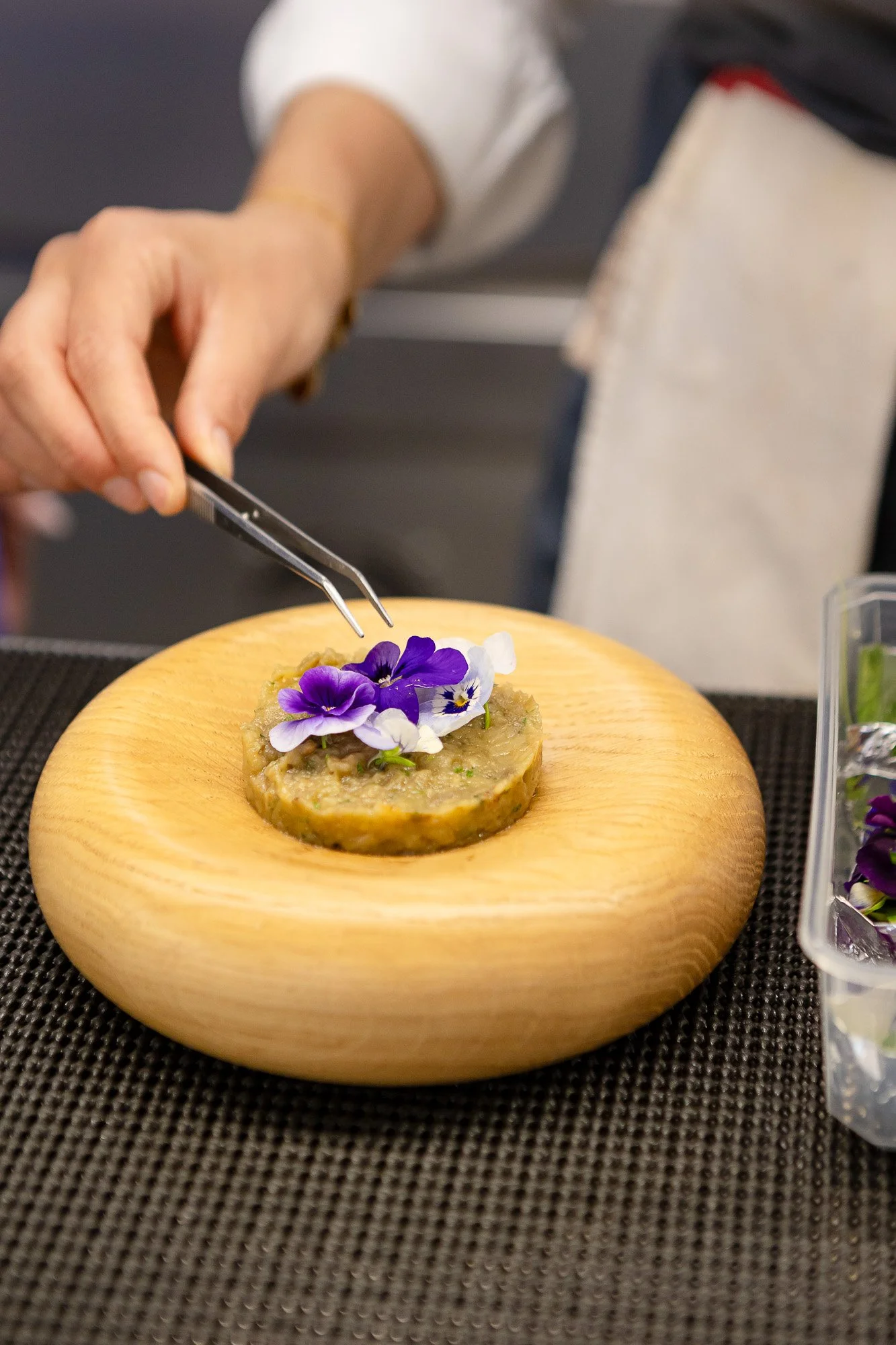 Une personne décore un plat avec des fleurs violettes et blanches sur une assiette en bois.