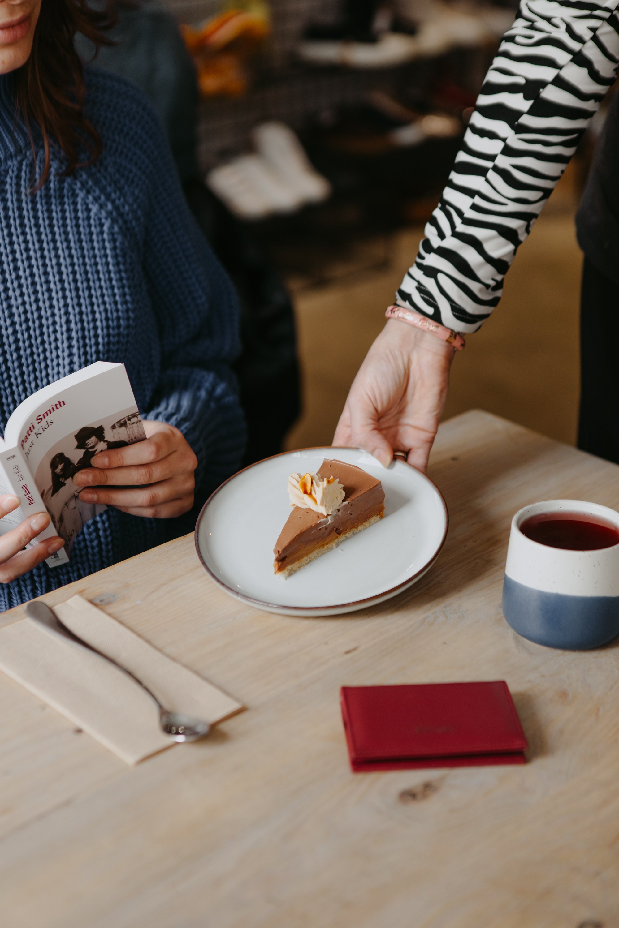 Une part de gâteau au chocolat sur une assiette, une tasse de boisson chaude, et une personne tendant la plaque à gâteau dans un café.
