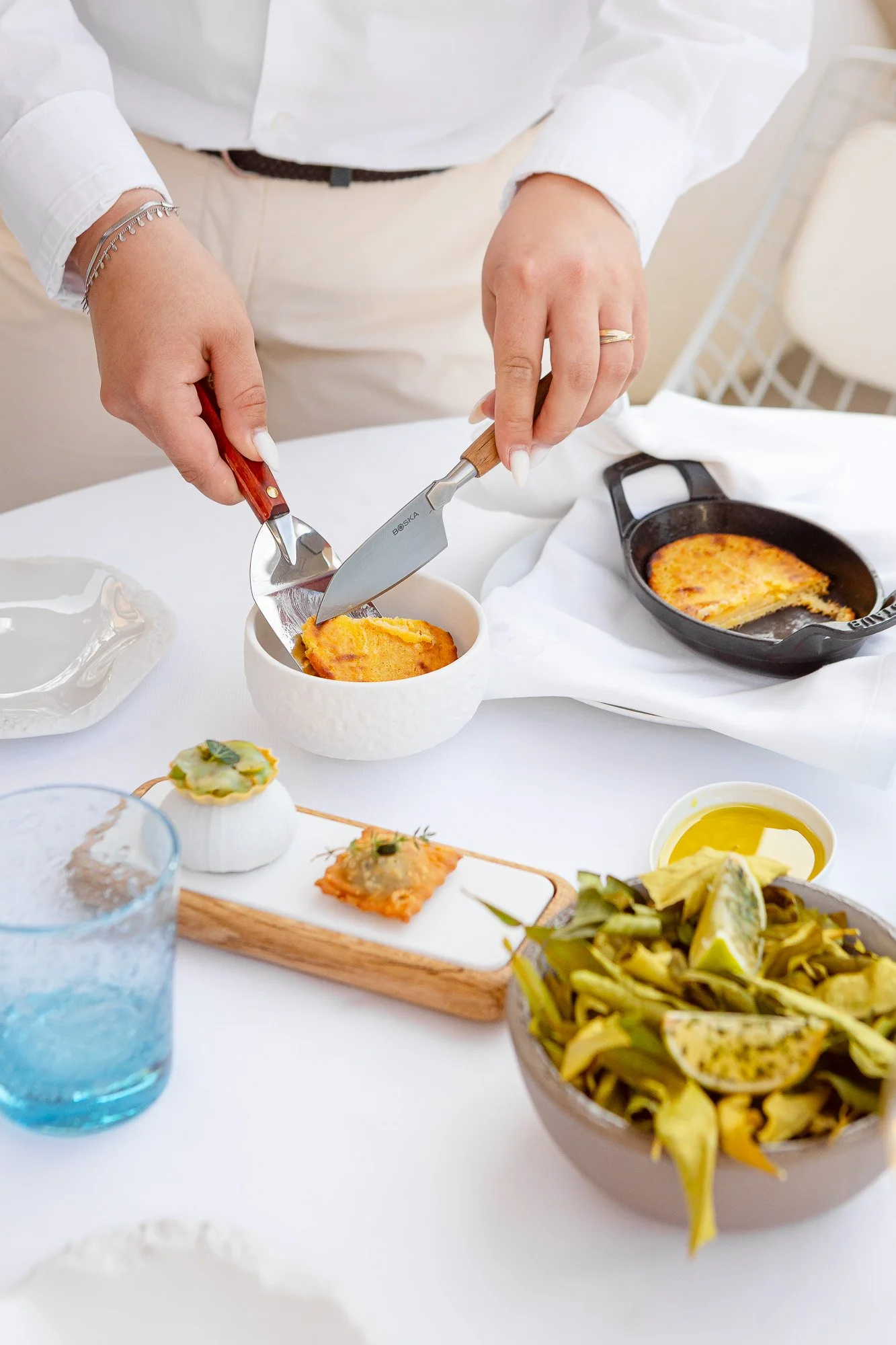Person coupant un quiche dans un bol blanc, entourée de diverses assiettes et plats avec salade, huiles et amuse-bouches, sur une table blanche, ambiance élégante.