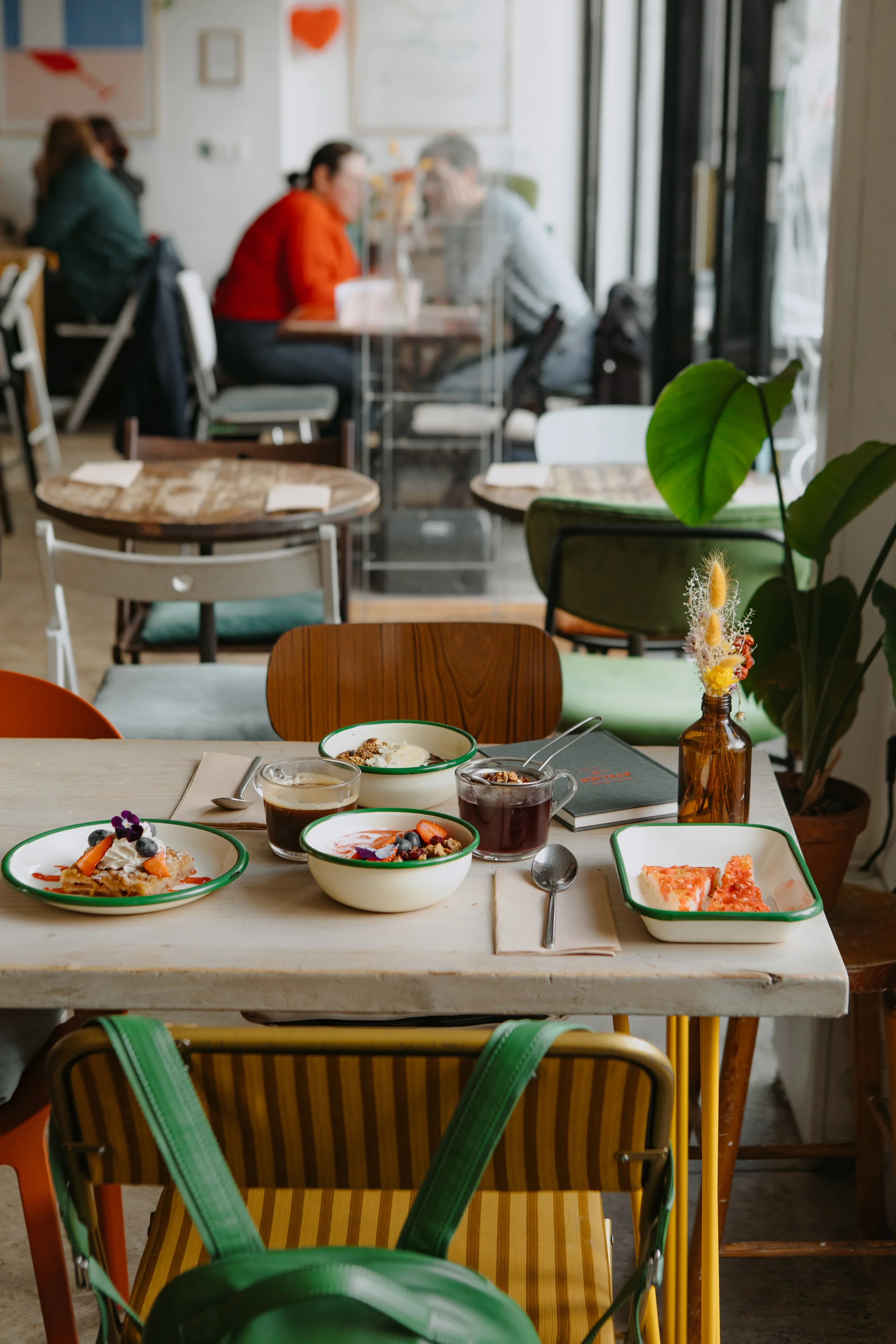 Une table dans un café ou restaurant avec des desserts et boissons, derrière une plante verte, plusieurs chaises colorées, et des personnes en arrière-plan à une autre table.
