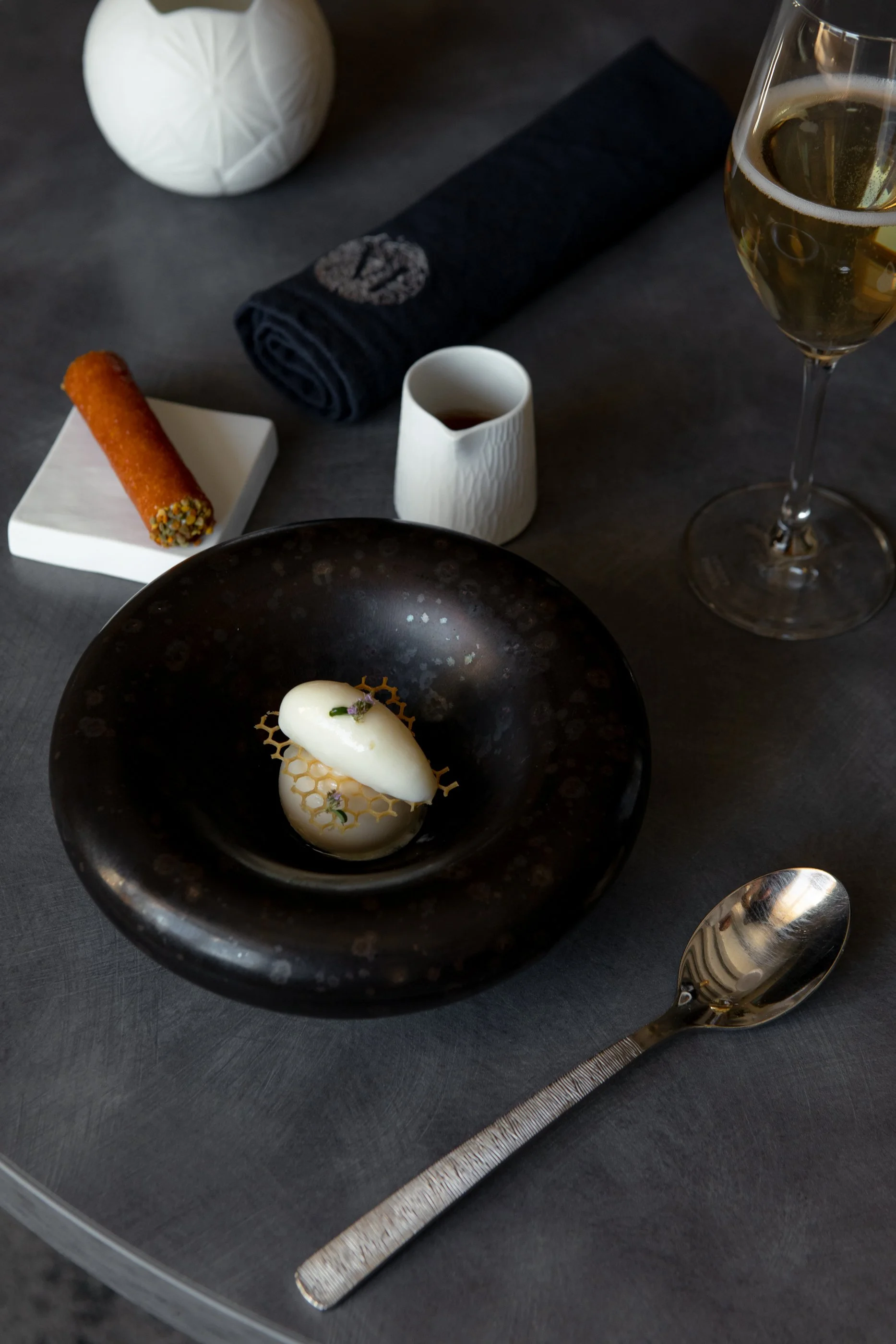 Un plat sophistiqué dans un bol noir avec une boule de glace ou de mousse blanche, décorée de motifs en crinoline, accompagné de verre de vin blanc et d'une cigarette russe croquante sur un plateau blanc. En arrière-plan, un vase blanc, un rouleau de
