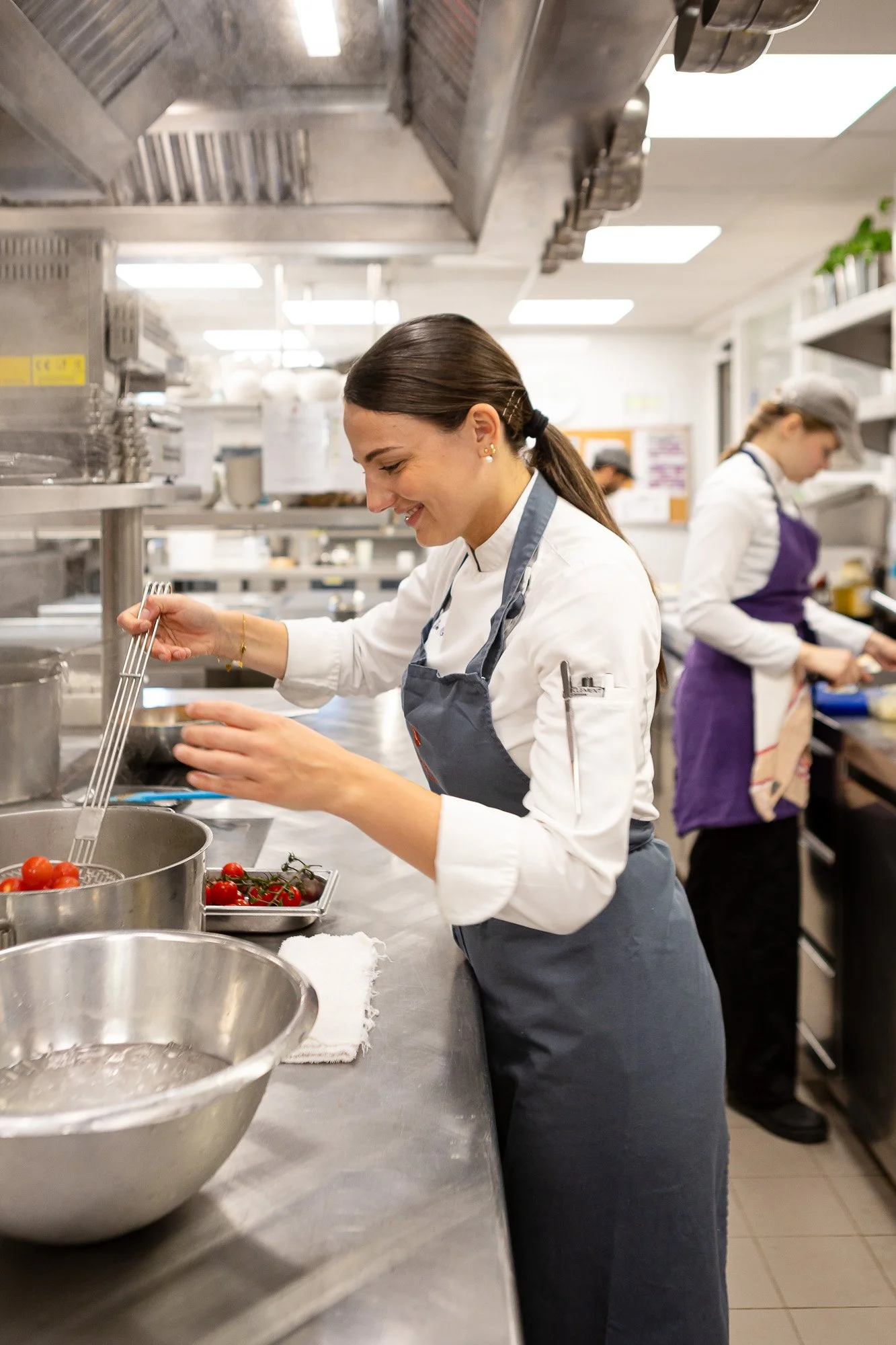 Une femme en Uniforme de chef cuisinier prépare des tomates dans une cuisine professionnelle, avec une femme en arrière-plan également en uniforme de cuisine.