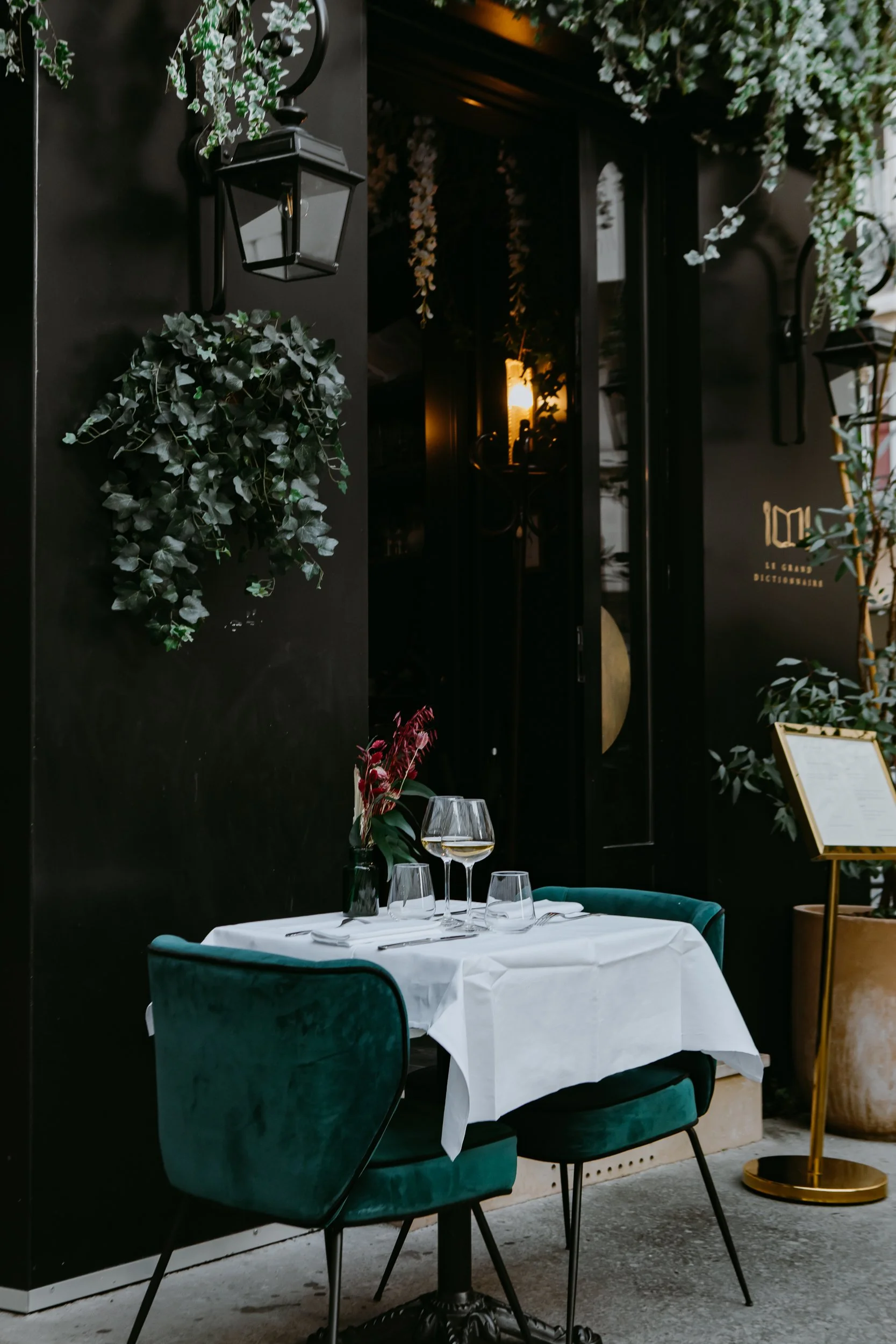 Une table de restaurant élégante avec une nappe blanche, des verres et des couverts, entourée de chaises en velours vert foncé, dans un décor chic avec des plantes et une lumière chaleureuse.