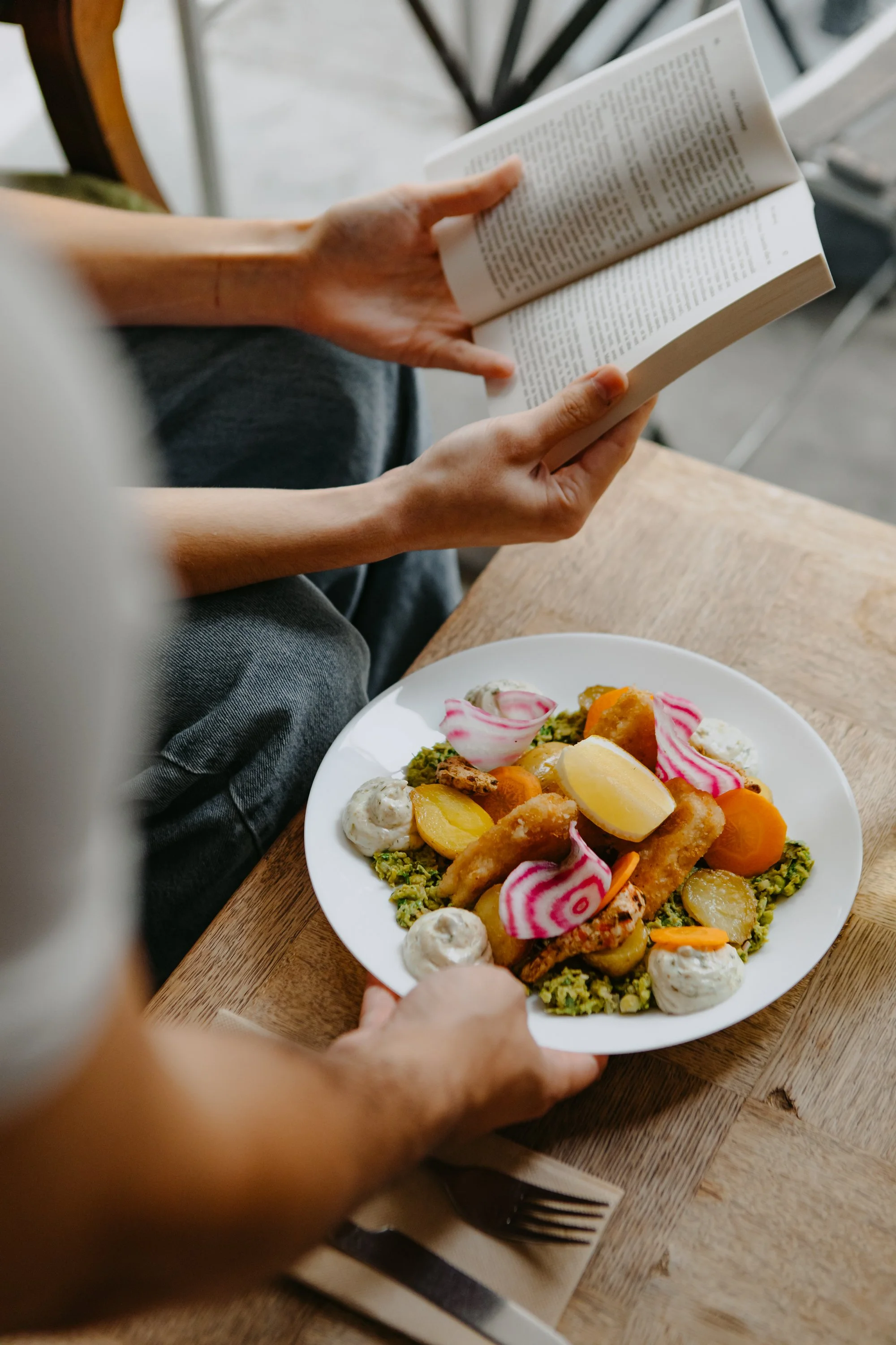 Une personne tient un livre ouvert, assise à une table en bois avec une assiette de nourriture colorée, comprenant des légumes, des morceaux de poisson ou de poulet, et des garnitures diverses.