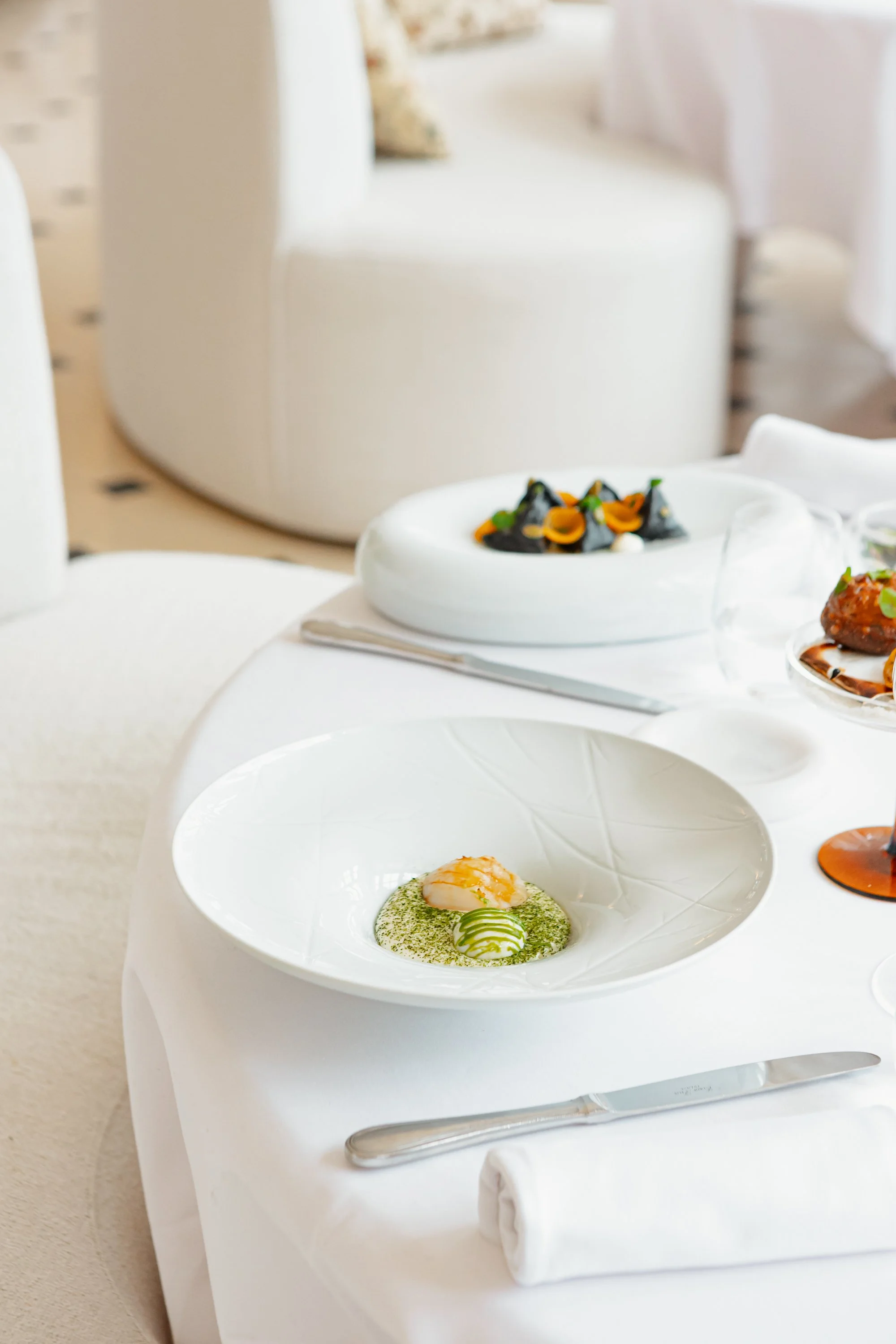 Plate de présentation gastronomique avec un œuf et une sauce verte, décorée de légumes roulés, sur une table blanche élégante avec des autres plats en arrière-plan.