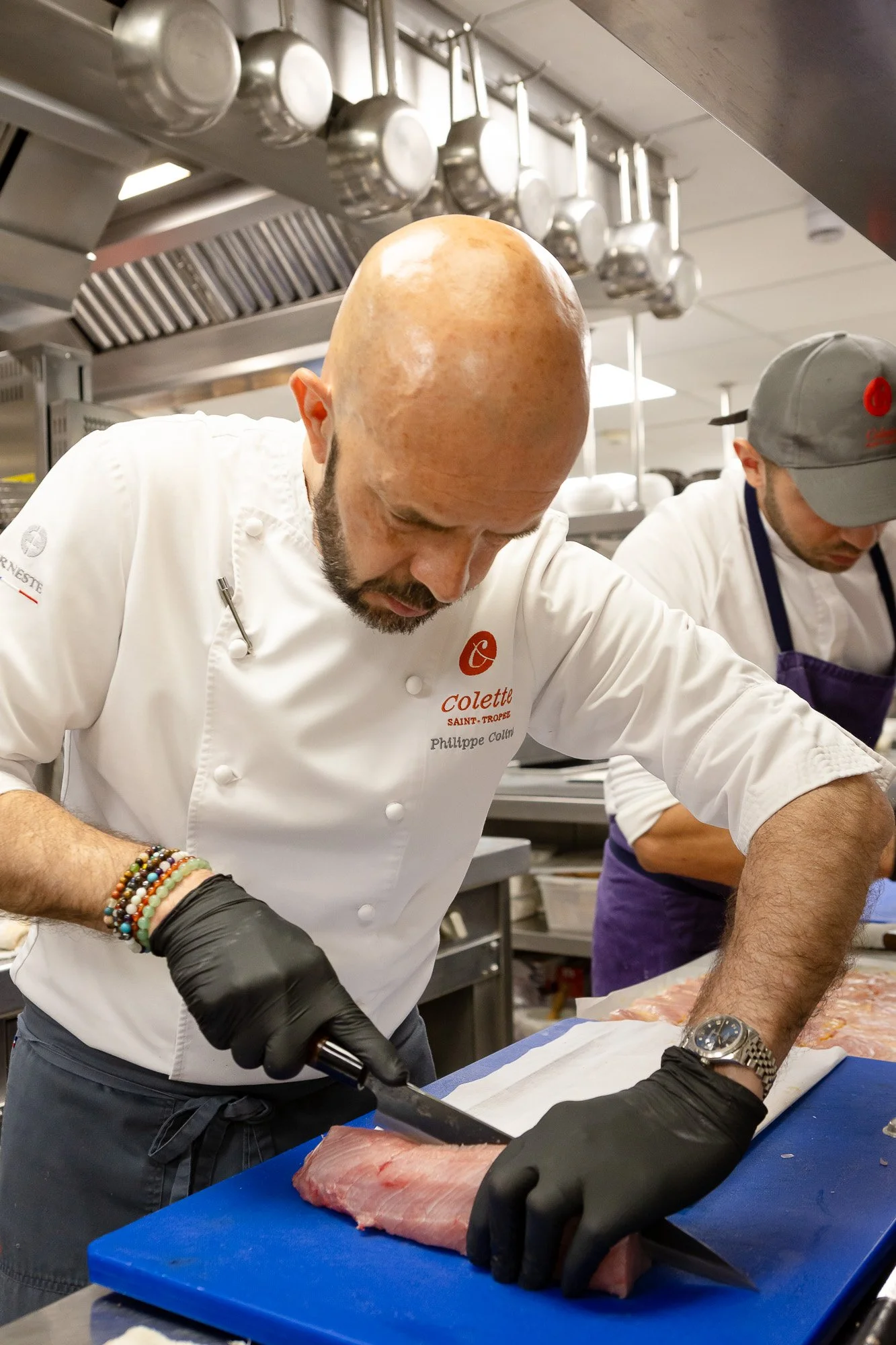 Un chef en costume blanc coupe un morceau de poisson sur une planche bleue, dans une cuisine professionnelle, avec un autre chef en arrière-plan.