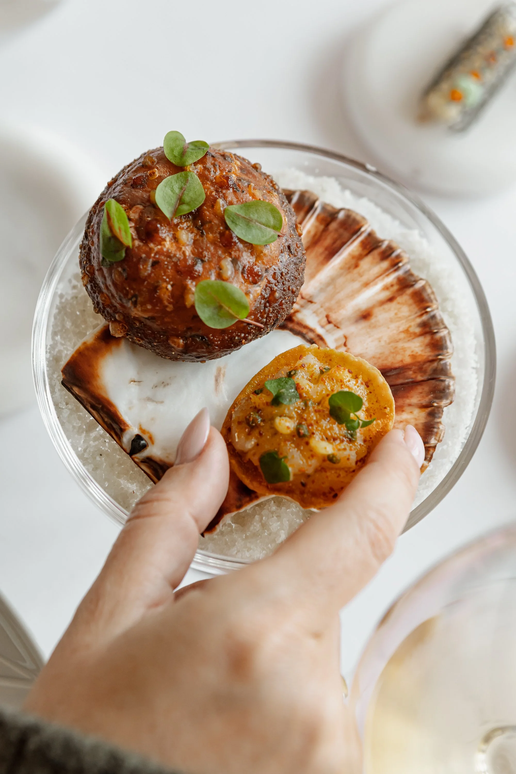 Une main tenant un plat avec des fruits de mer, notamment un coquillage ovale et une boule de viande, tous garnis de petites feuilles vertes.