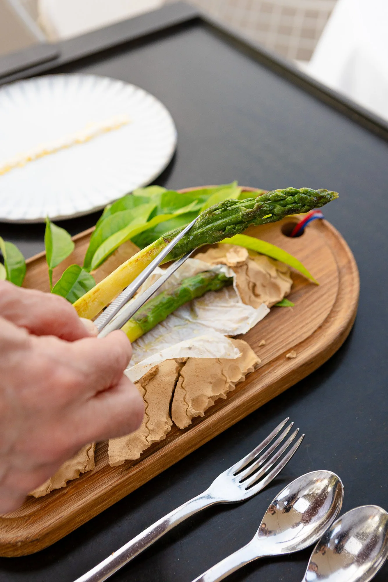 Une personne utilise des pinces pour saisir un asperge verte sur un plateau en bois avec du fromage à pâte molle, des feuilles vertes et une assiette blanche en arrière-plan.