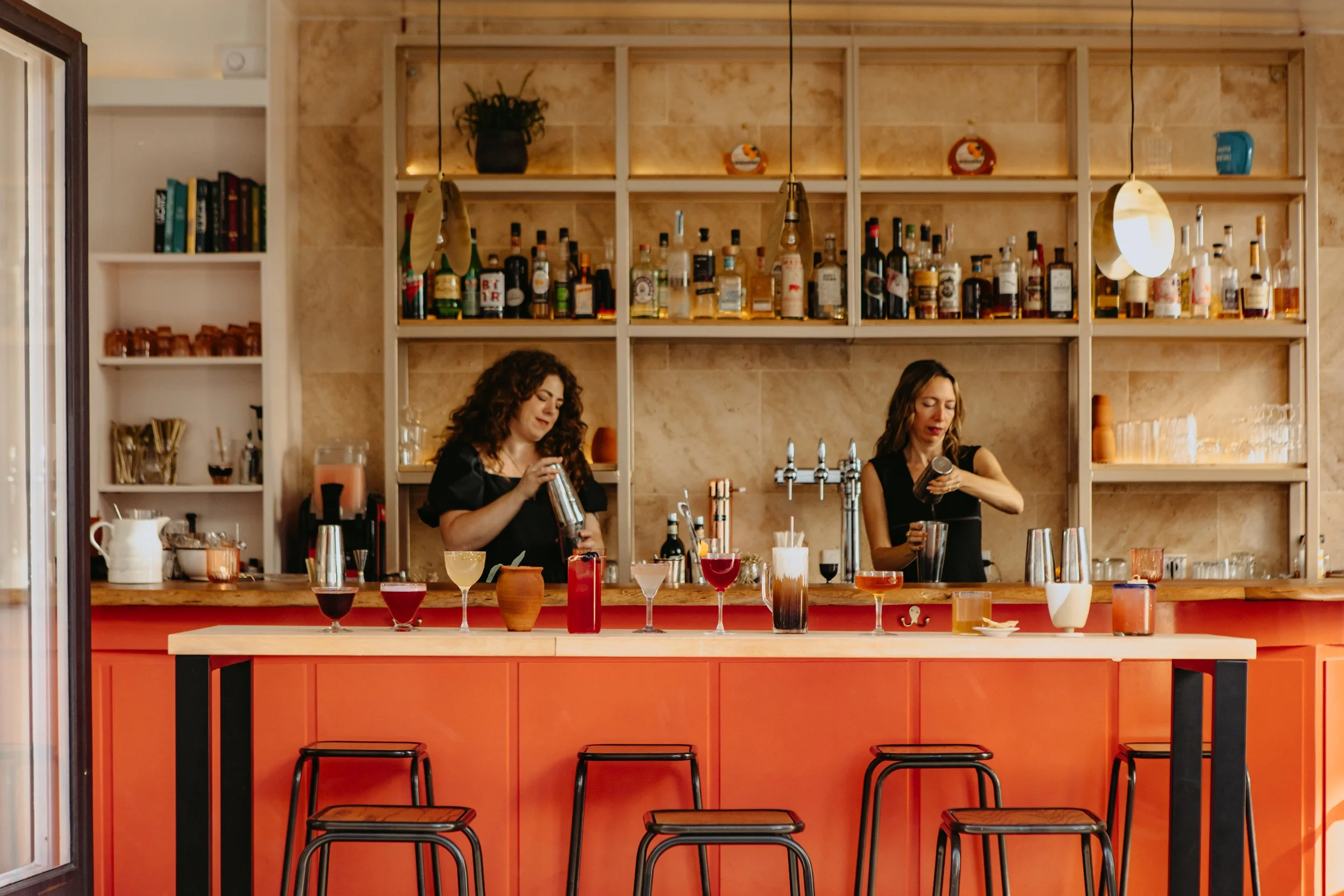 Deux femmes préparent des cocktails derrière un bar orange avec des tabourets noirs, des bouteilles d'alcool sur des étagères en arrière-plan et des verres colorés sur le comptoir.