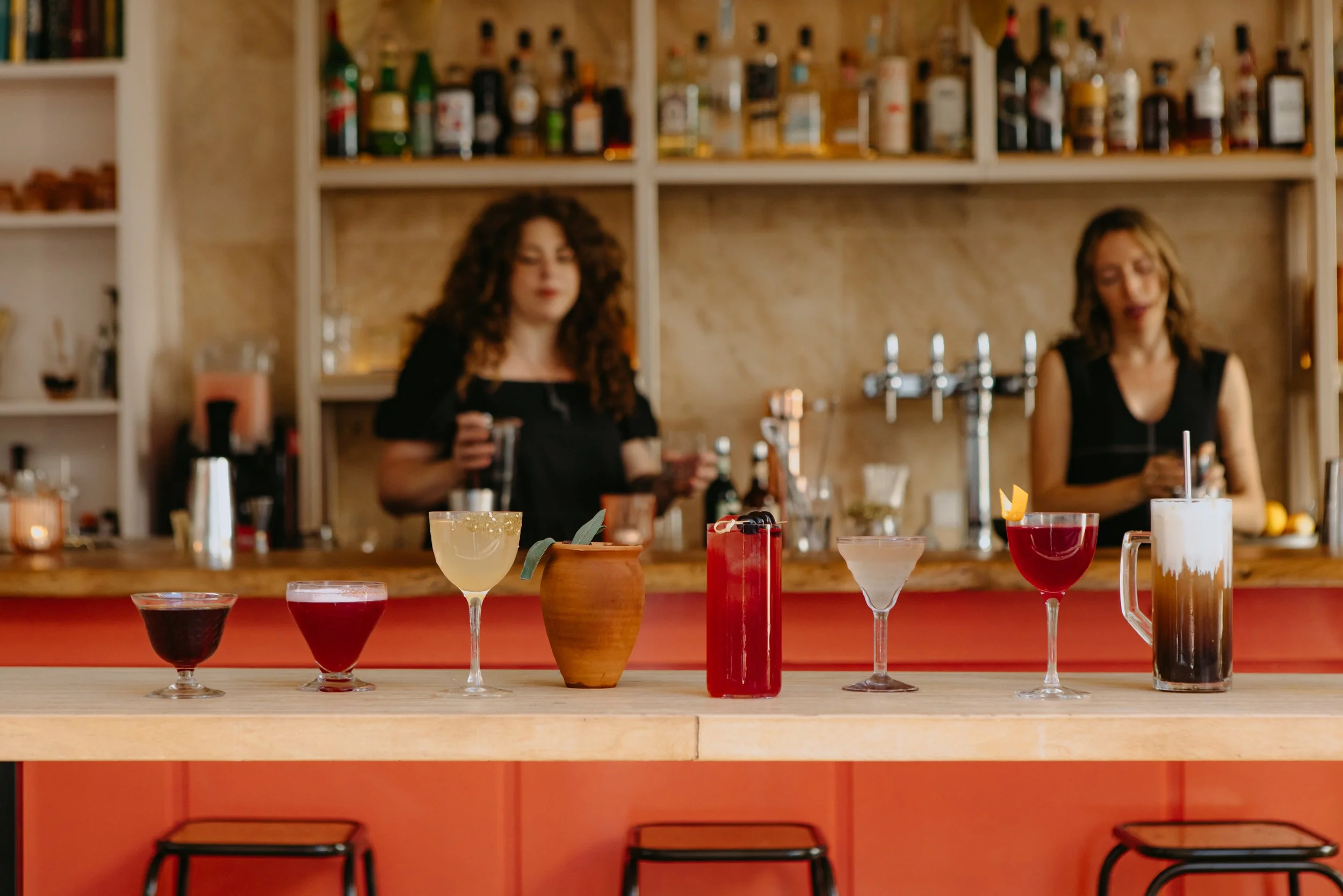 Une bar avec différentes boissons colorées alignées sur le comptoir, deux femmes avec des verres en main en arrière-plan, dans un décor de bar chaleureux et accueillant.