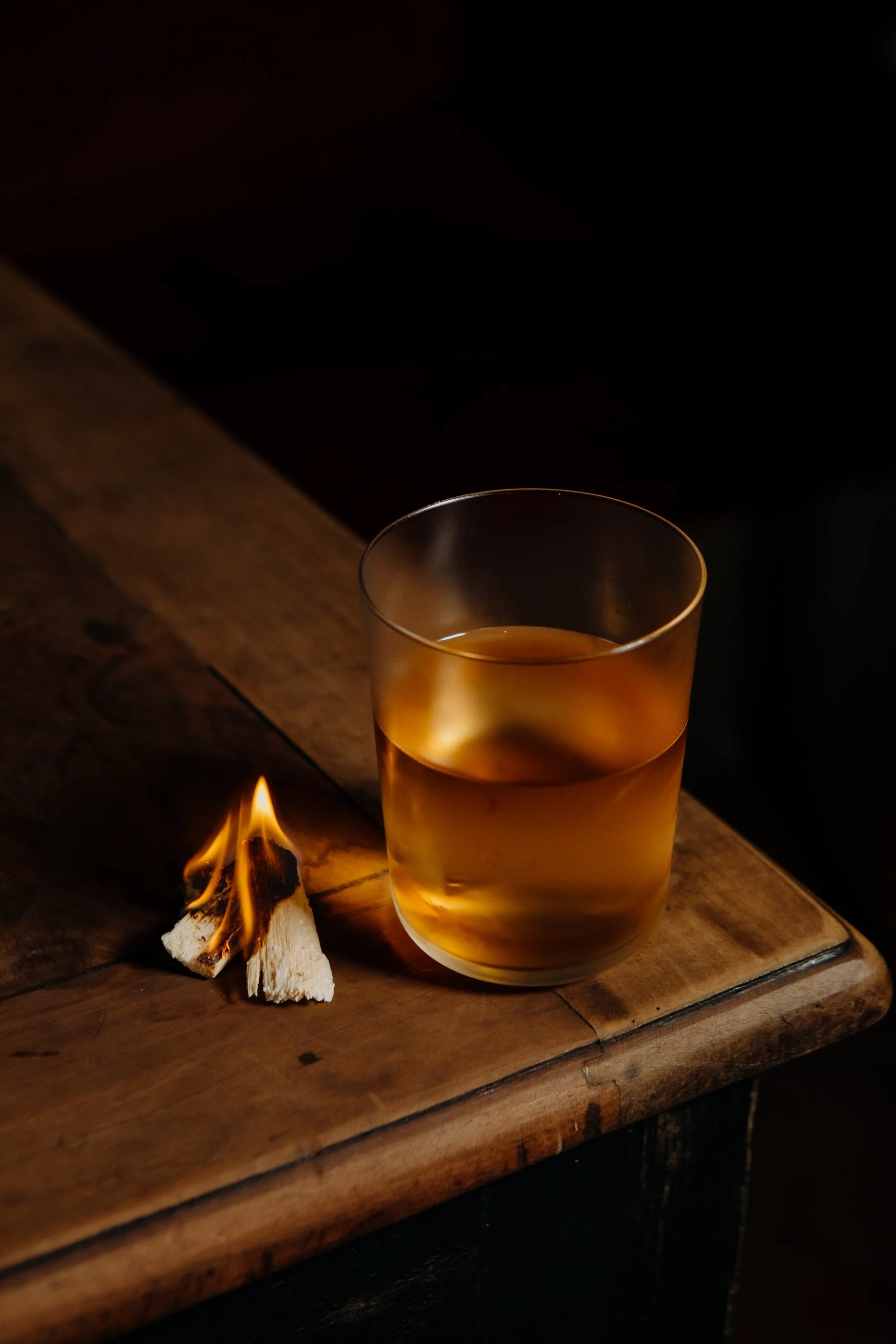 Verre avec une boisson dorée sur une table en bois, avec un petit feu de bois allumé à côté.