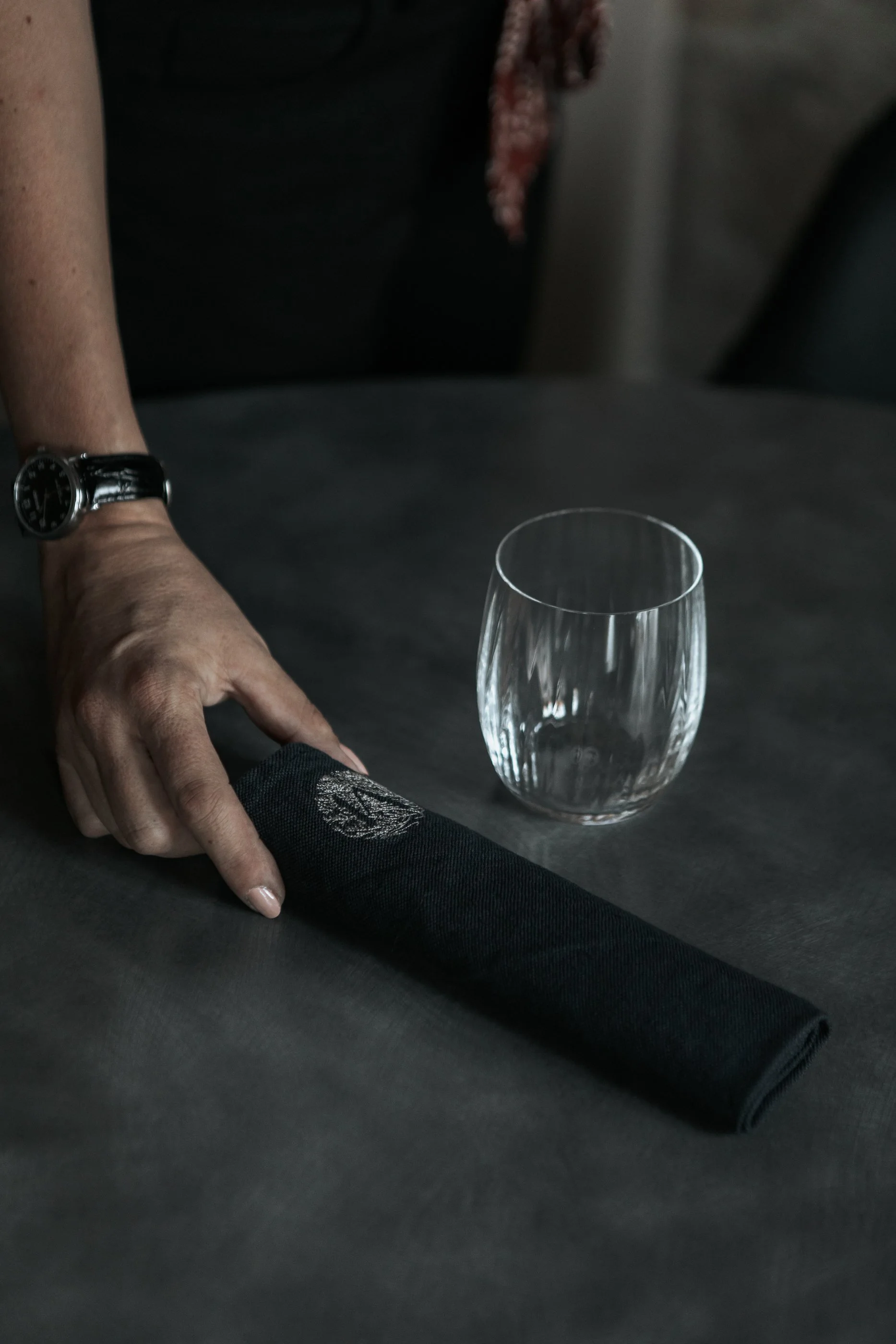 Une main posée sur une nappe noire, un verre en cristal vide à côté.