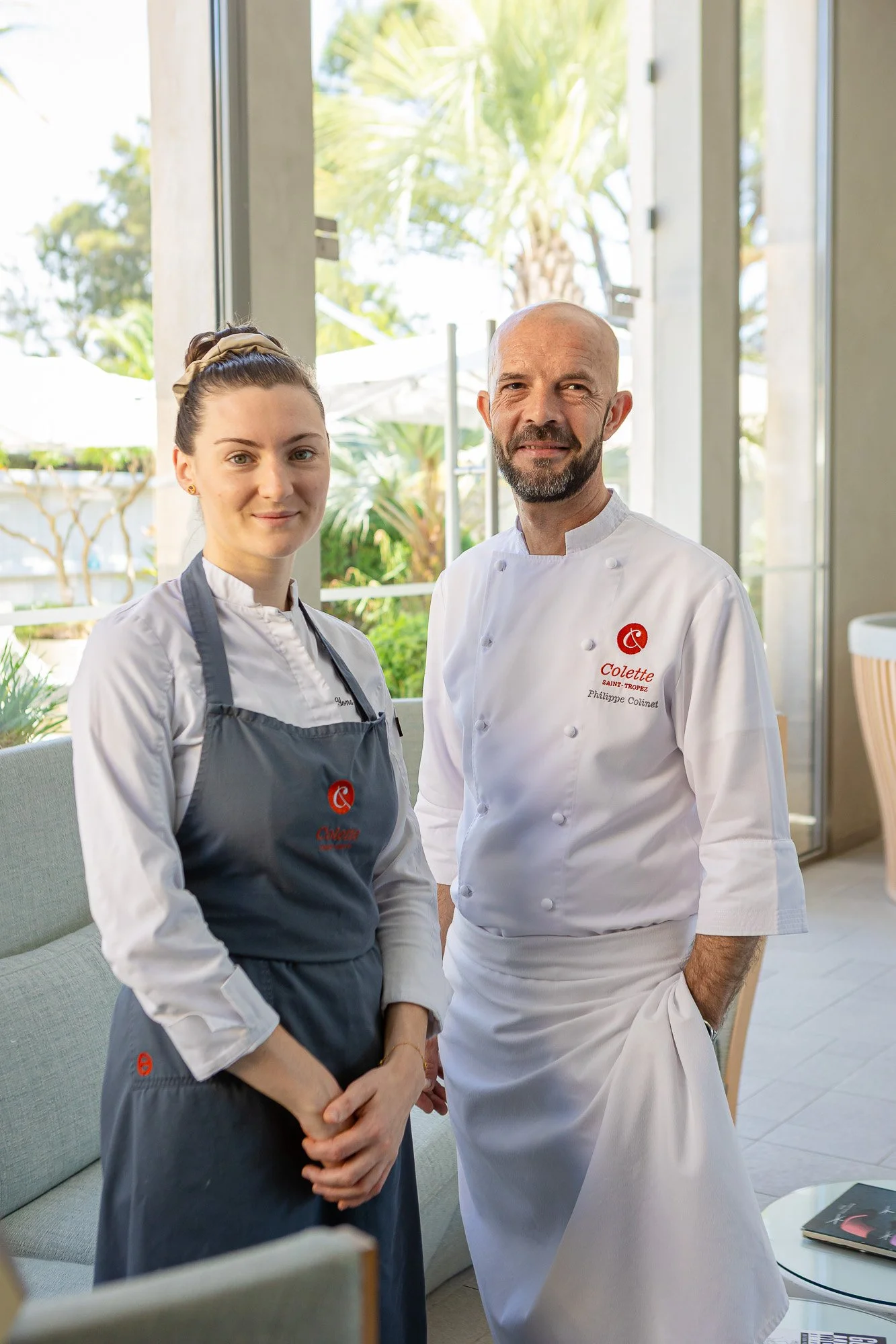 Deux chefs en uniforme posent dans une cuisine ensoleillée avec des vitres grandes ouvertes, des plantes tropicales et une vue sur l'extérieur.
