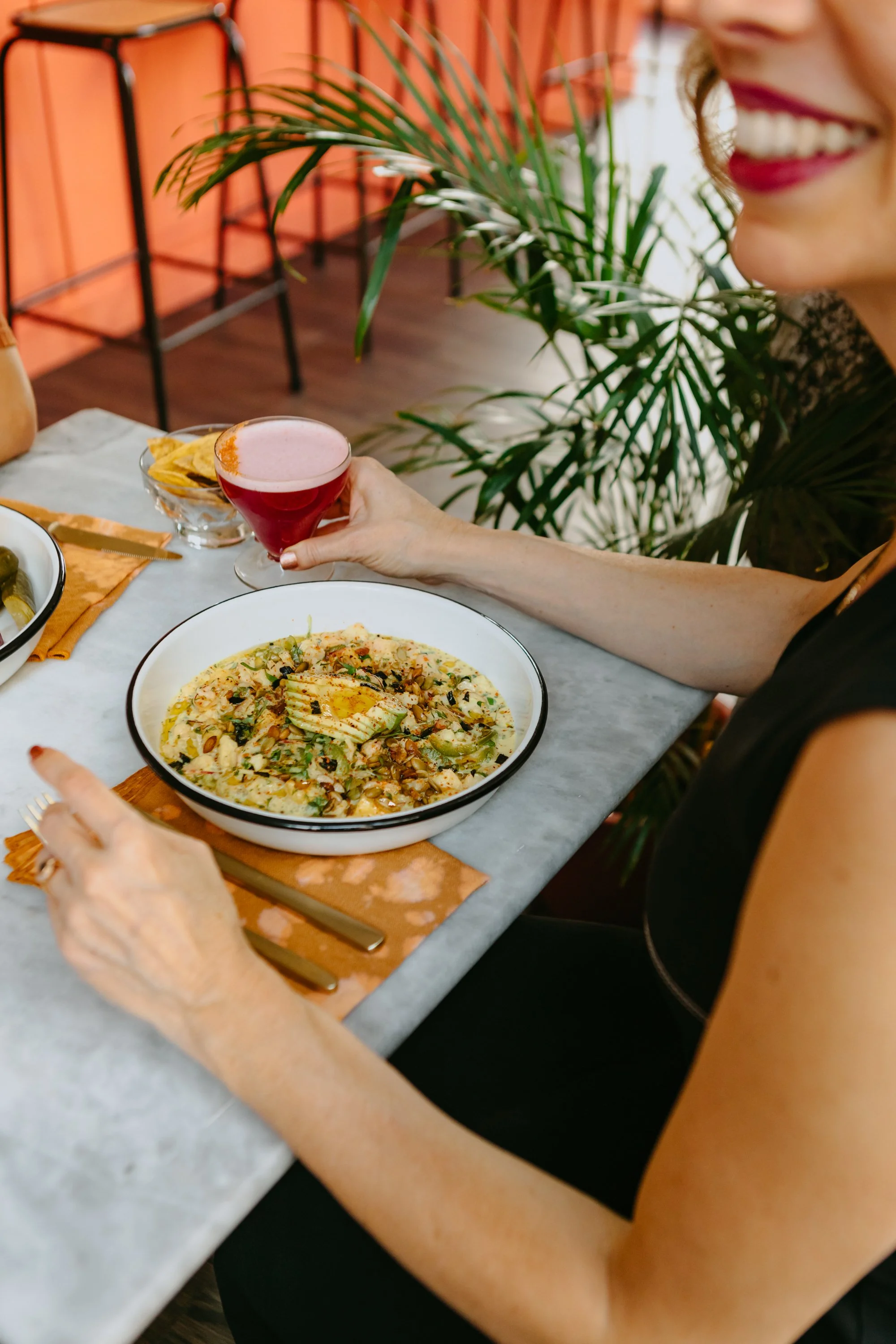 Une femme souriante, assise à une table, tenant un cocktail rouge entourée de plats et d'accessoires de table dans un décor intérieur avec des plantes vertes.